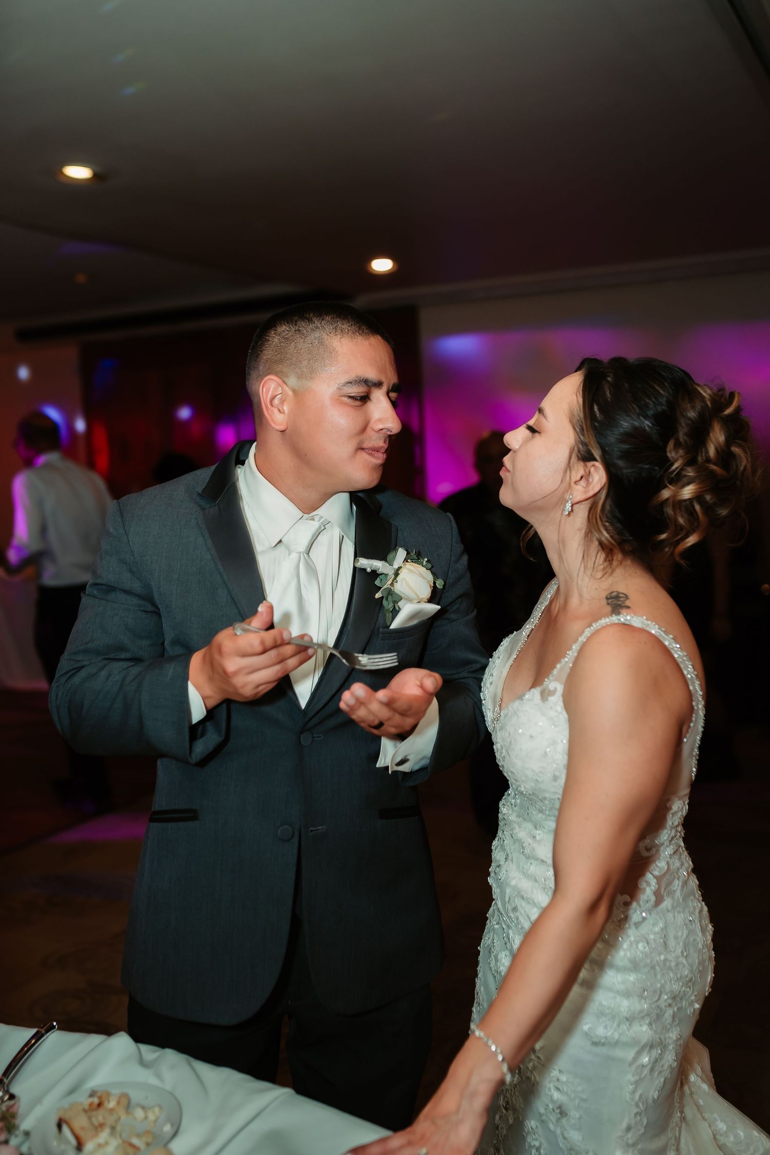 Newlyweds playfully feeding each other wedding cake during reception celebration.