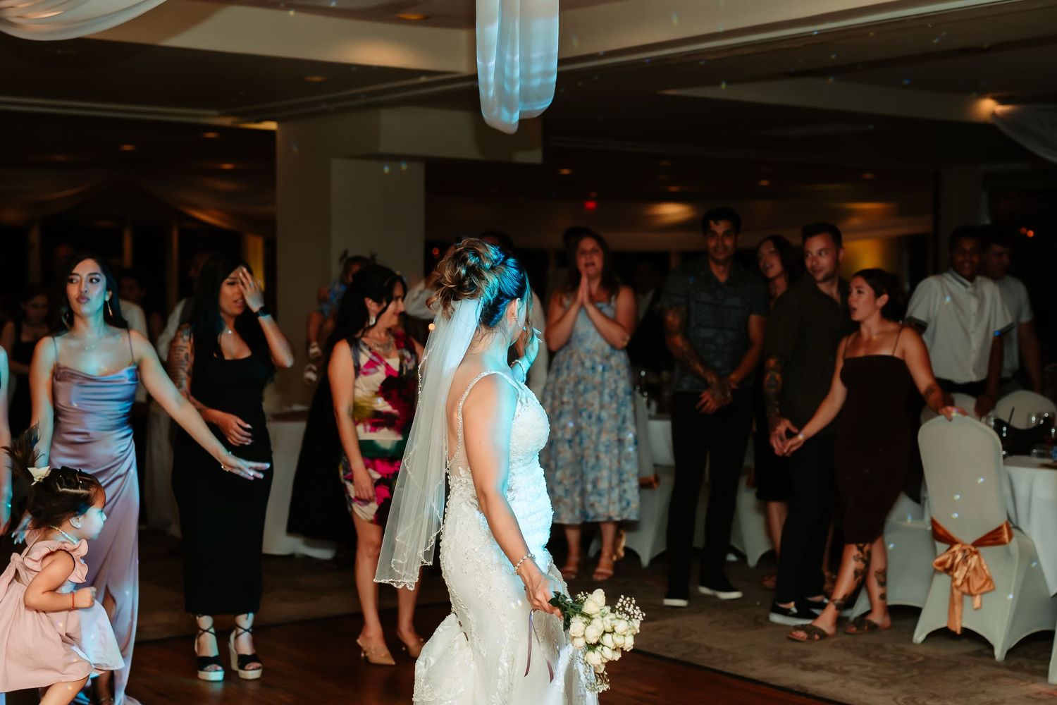A bride in white dress tosses her bouquet to waiting guests at a wedding reception venue with blue lighting.