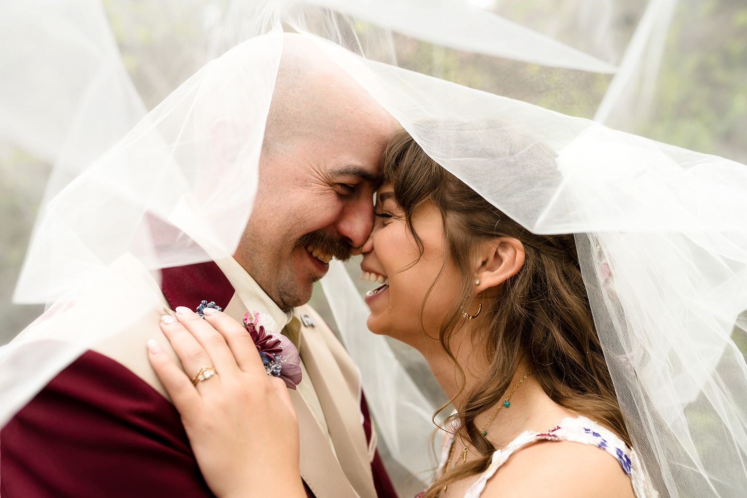 Bride and groom smiling at each other under a wedding veil in an intimate romantic portrait.