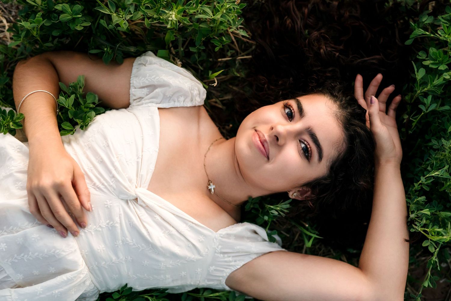 Young woman in white dress lying on green grass, looking up with dark curly hair and delicate necklace.
