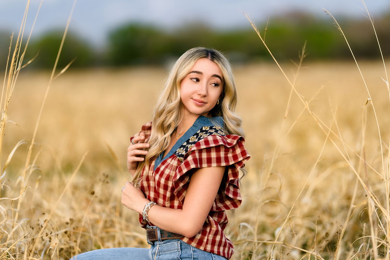 Young blonde woman in plaid shirt poses in golden wheat field, holding her hair while looking over shoulder.