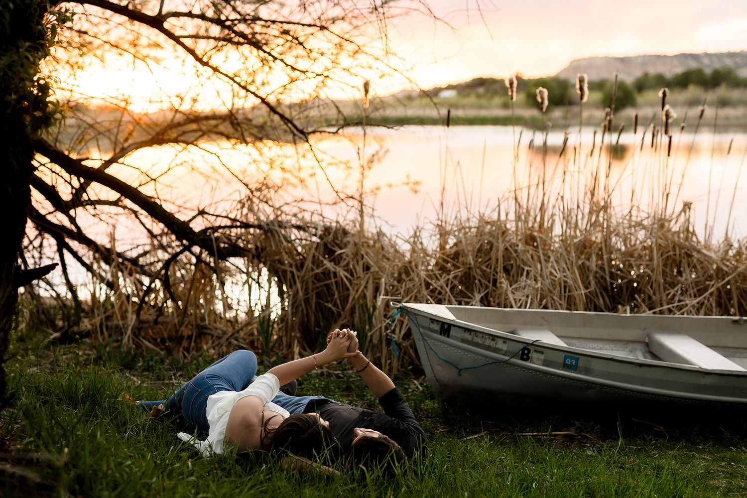 Person lying on grass by a calm lake at sunset near a rowboat, surrounded by reeds and trees.