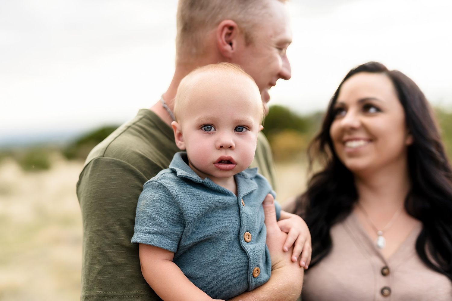 A family portrait of a man holding a baby boy in a blue outfit while a smiling woman looks on outdoors.