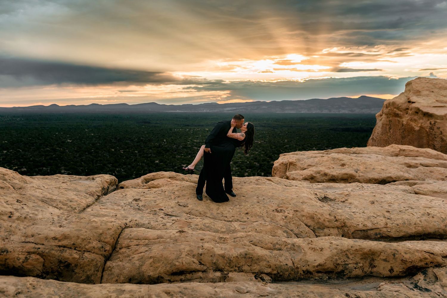 A couple in black embraces on a rocky cliff at sunset with a dramatic sky and vast landscape behind them.