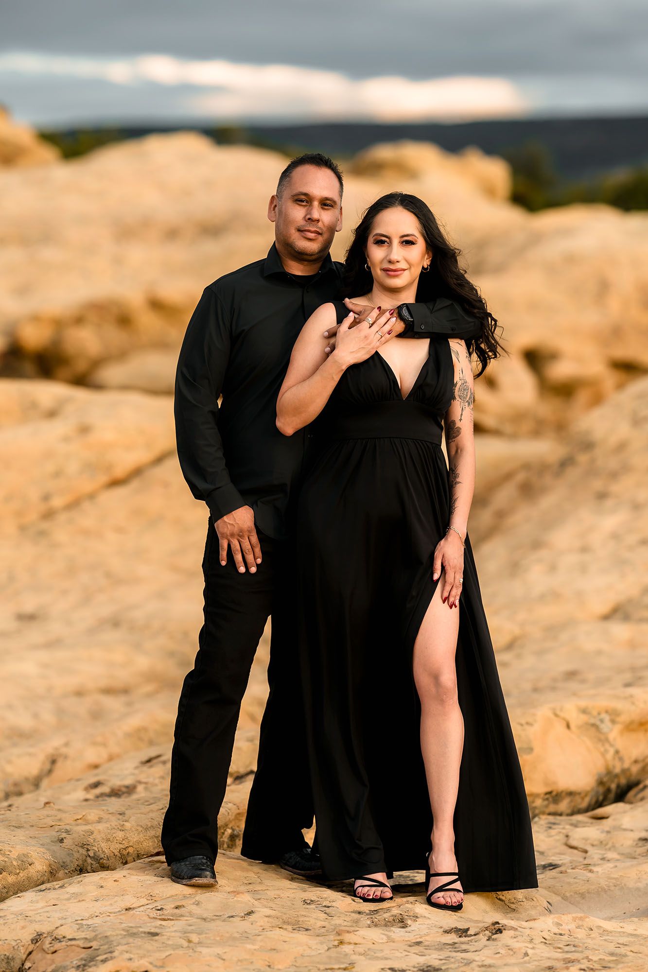 Elegant couple posing in matching black outfits on desert rocks at golden hour, woman in slit gown.