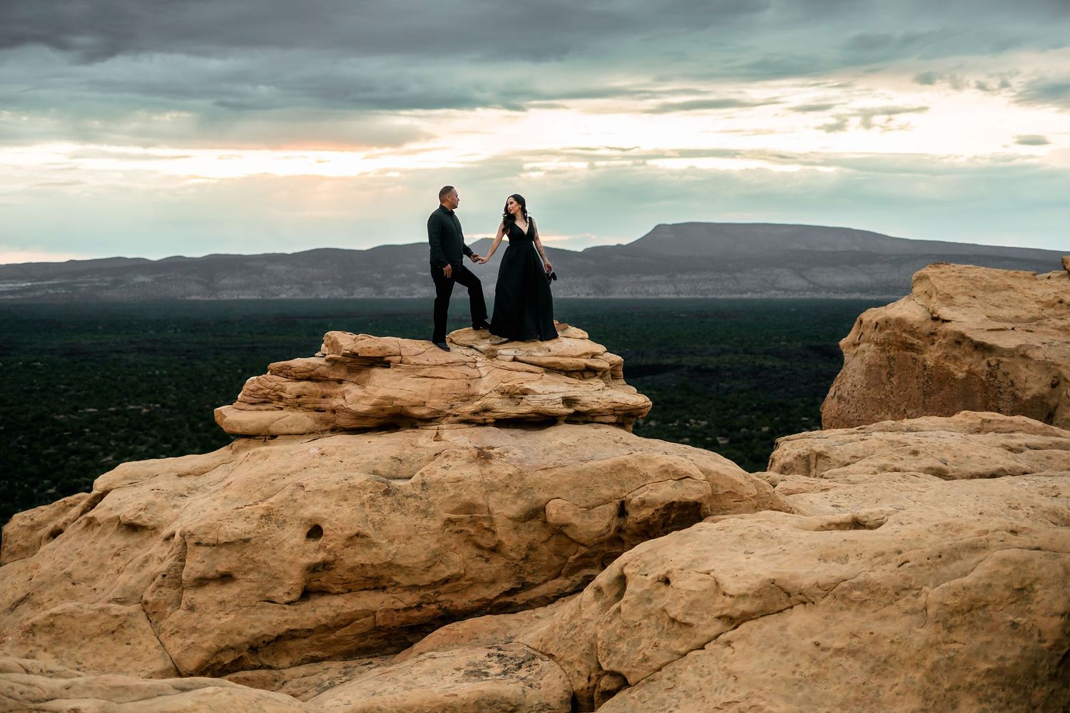 A couple in black formal attire holds hands on rocky cliffs with a dramatic cloudy sky backdrop.