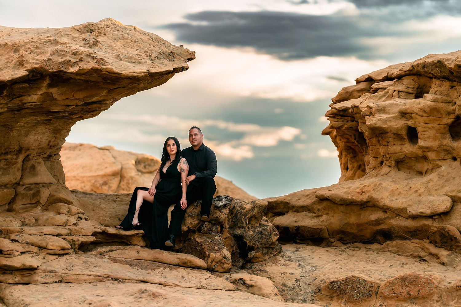Couple in black outfits sitting among dramatic sandstone rock formations under a cloudy sky.