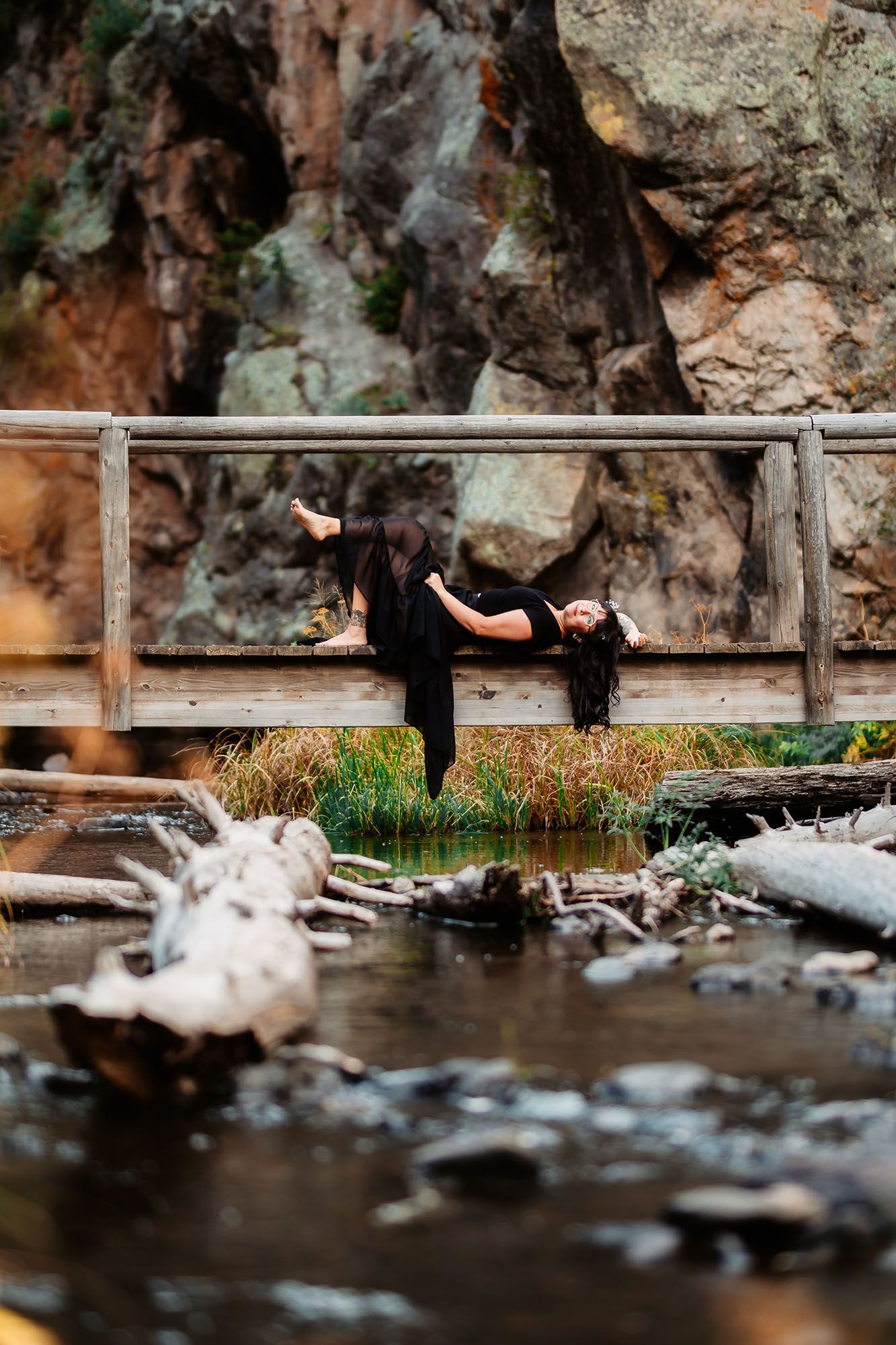 Woman in black outfit performing dramatic pose on wooden bridge over rocky mountain stream.