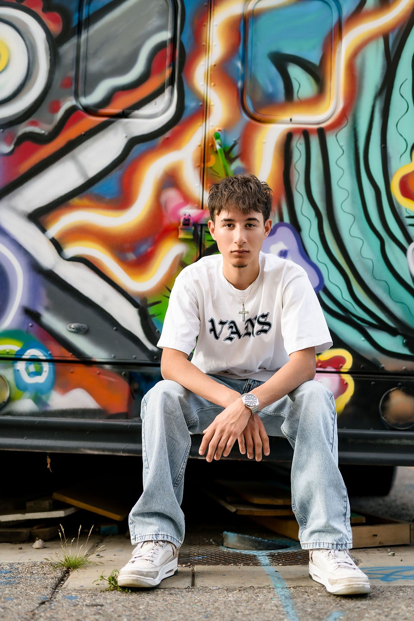 Teen boy in white Vans tee and jeans sits confidently in front of colorful graffiti wall.