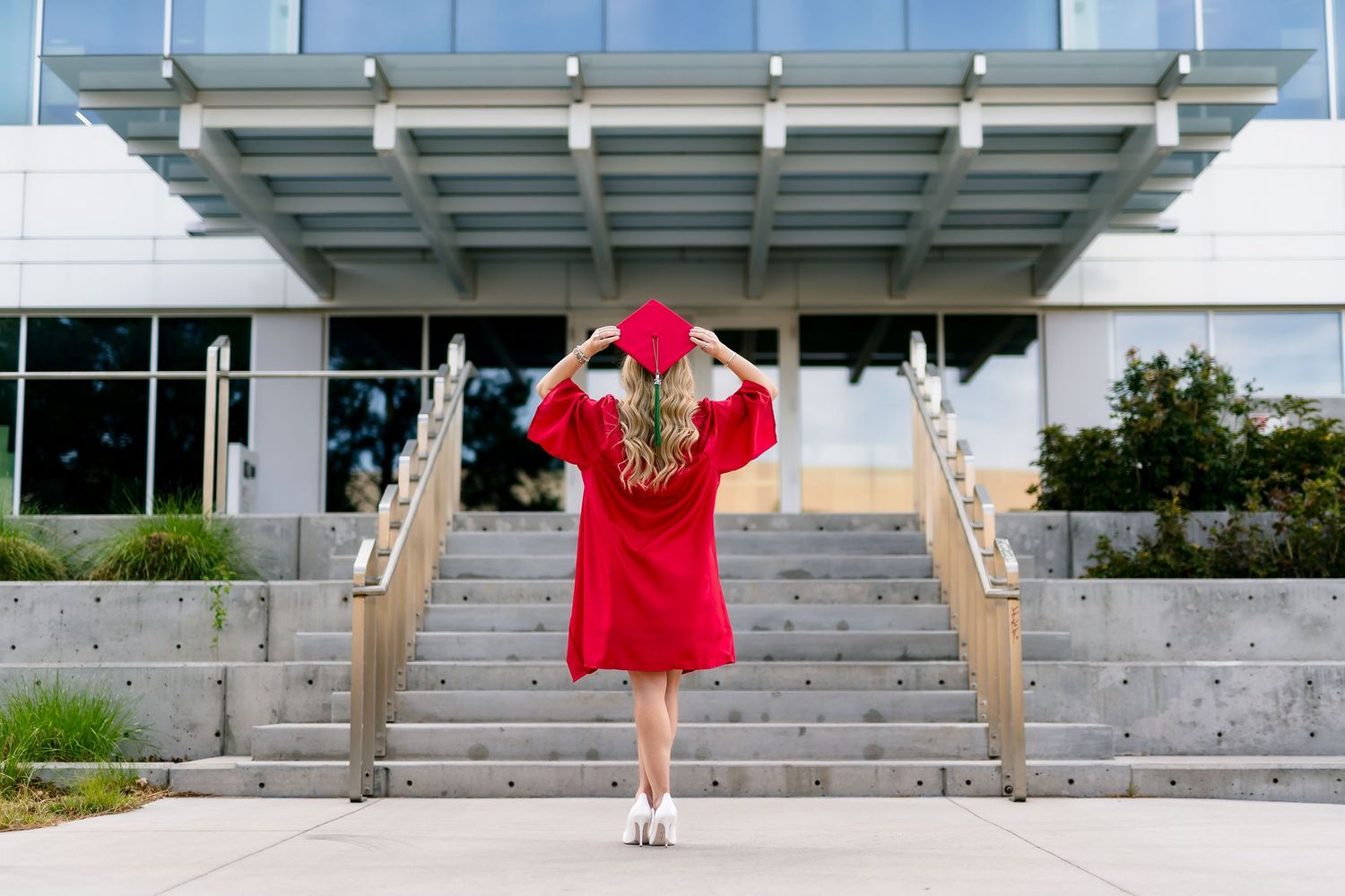 A blonde woman in a red graduation gown and cap stands on steps outside a modern building.