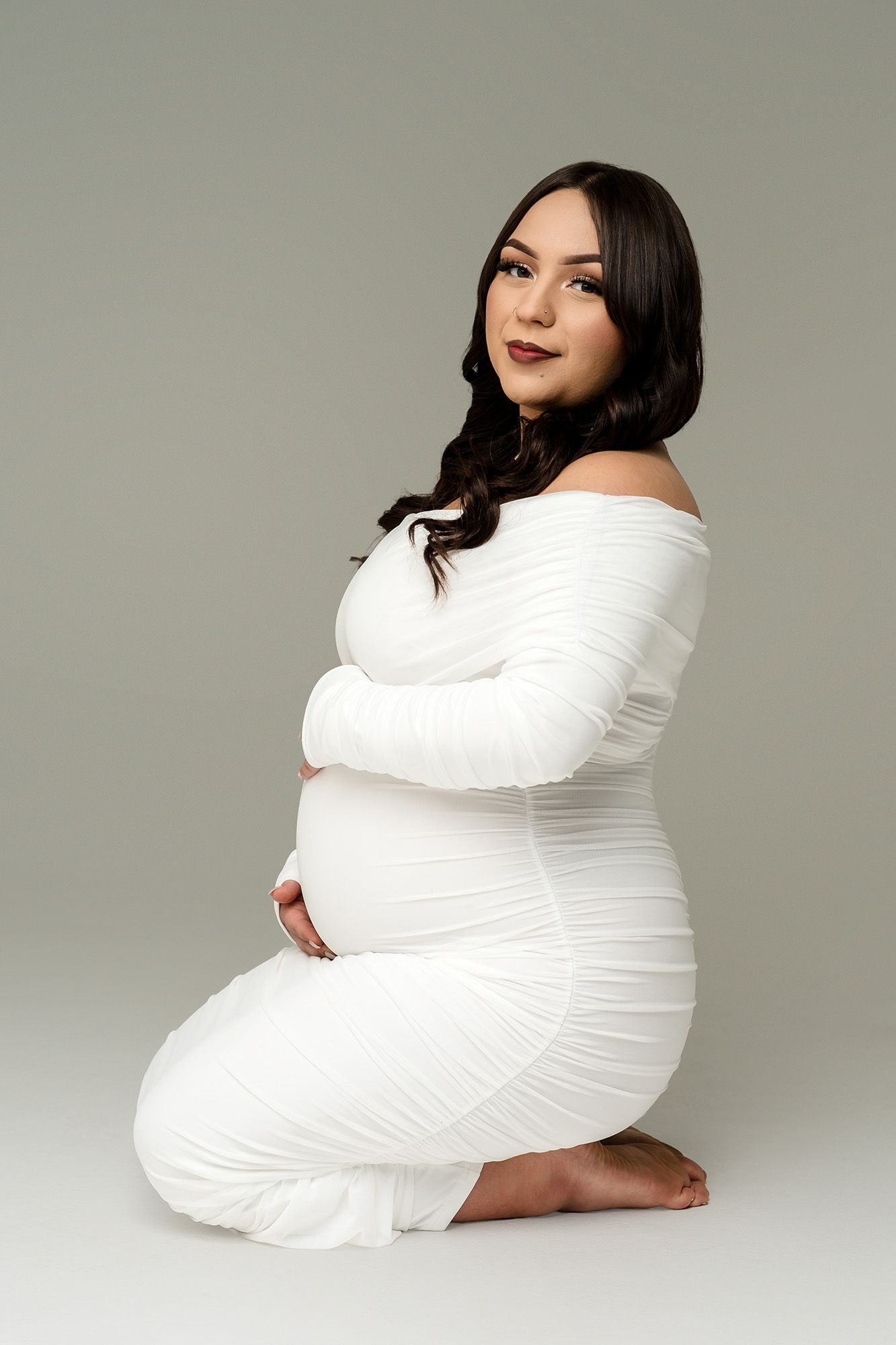 Pregnant woman in white off-shoulder ruched dress kneeling gracefully, cradling baby bump on gray background.