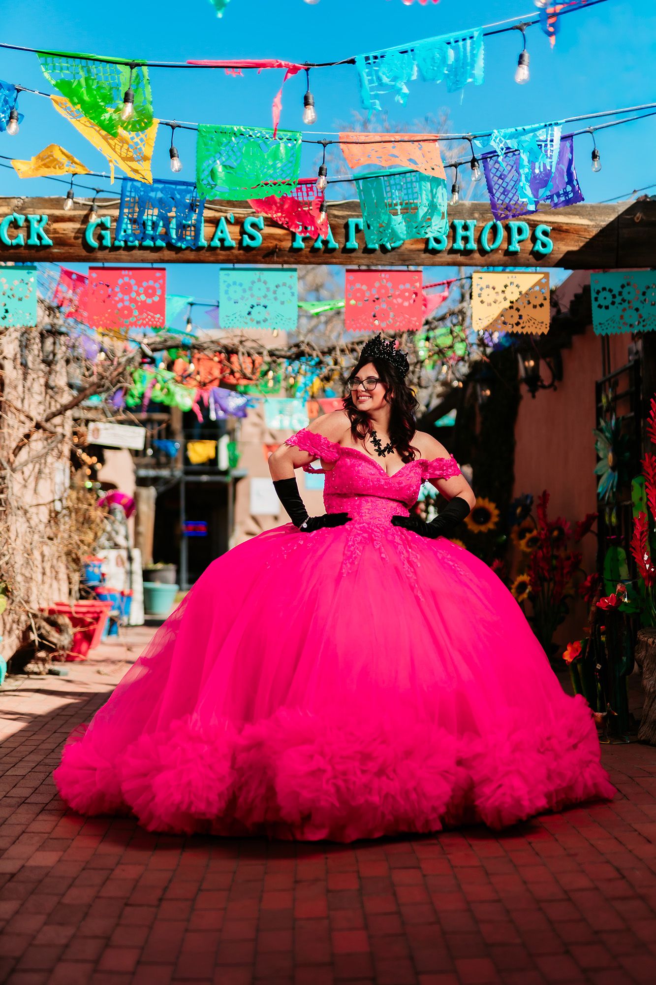 A bright pink quinceañera dress flows dramatically across a brick courtyard under colorful papel picado decorations.