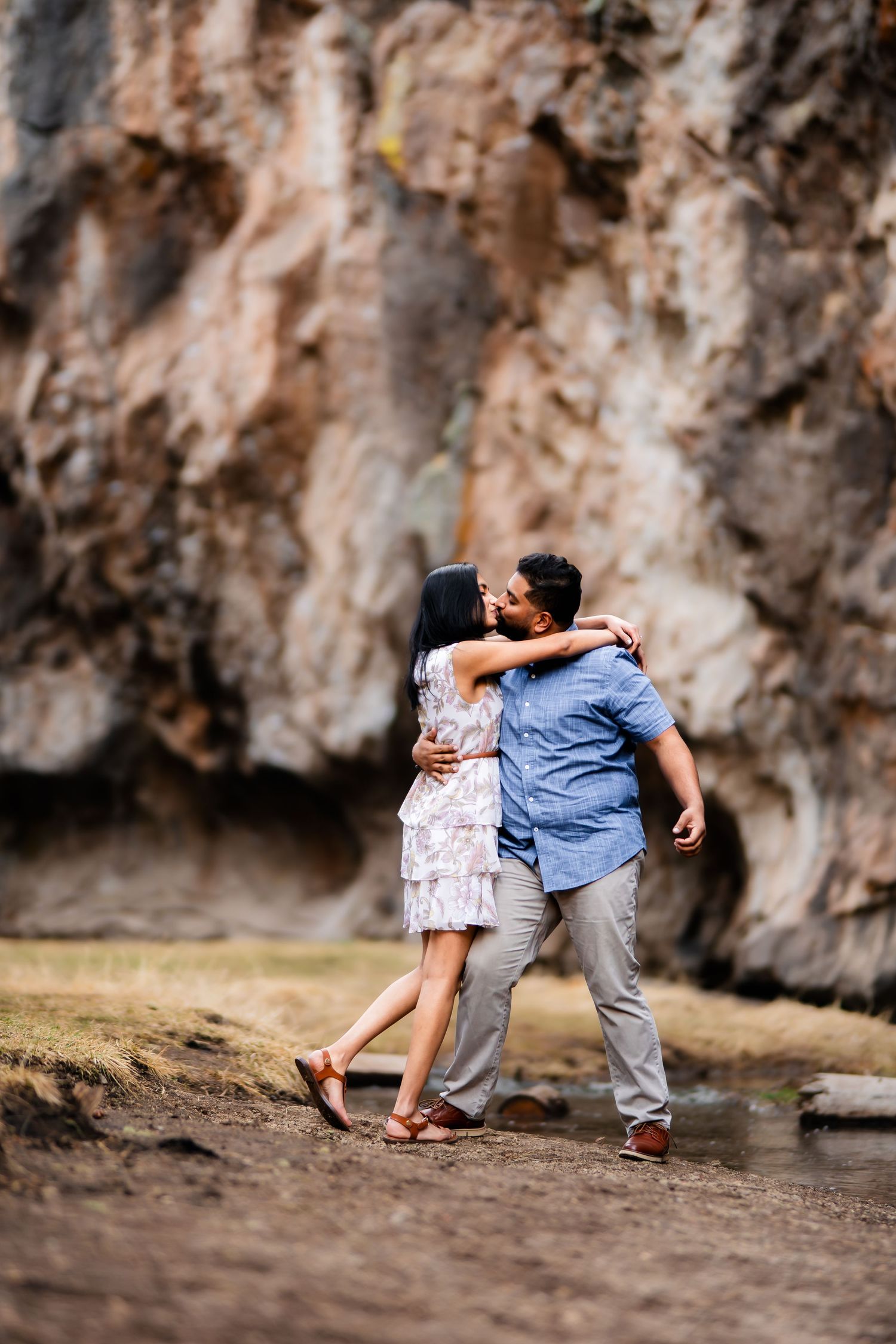 Couple embraces on trail beside rocky cliff face near mountain stream.
