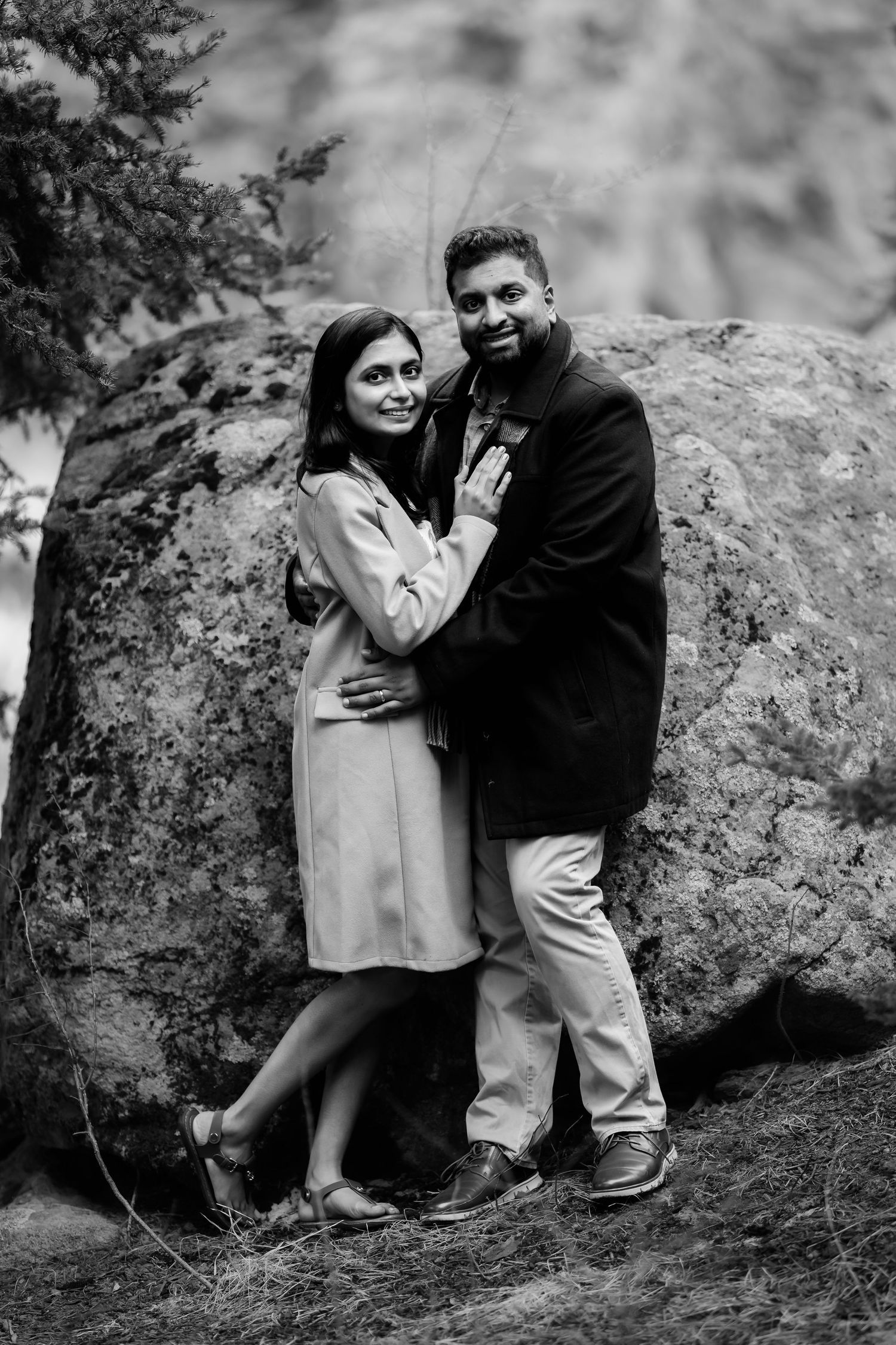 A couple in black and white embraces against a large boulder in a natural outdoor setting.