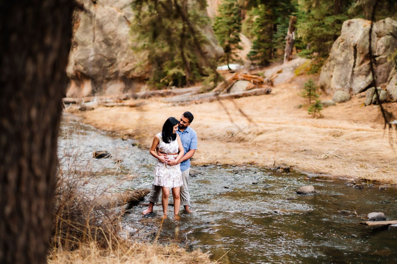 A couple stands together in shallow water surrounded by rocks and trees in a scenic mountain stream.
