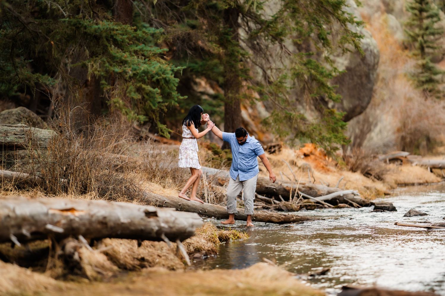A couple holds hands while walking along fallen logs by a scenic mountain stream.