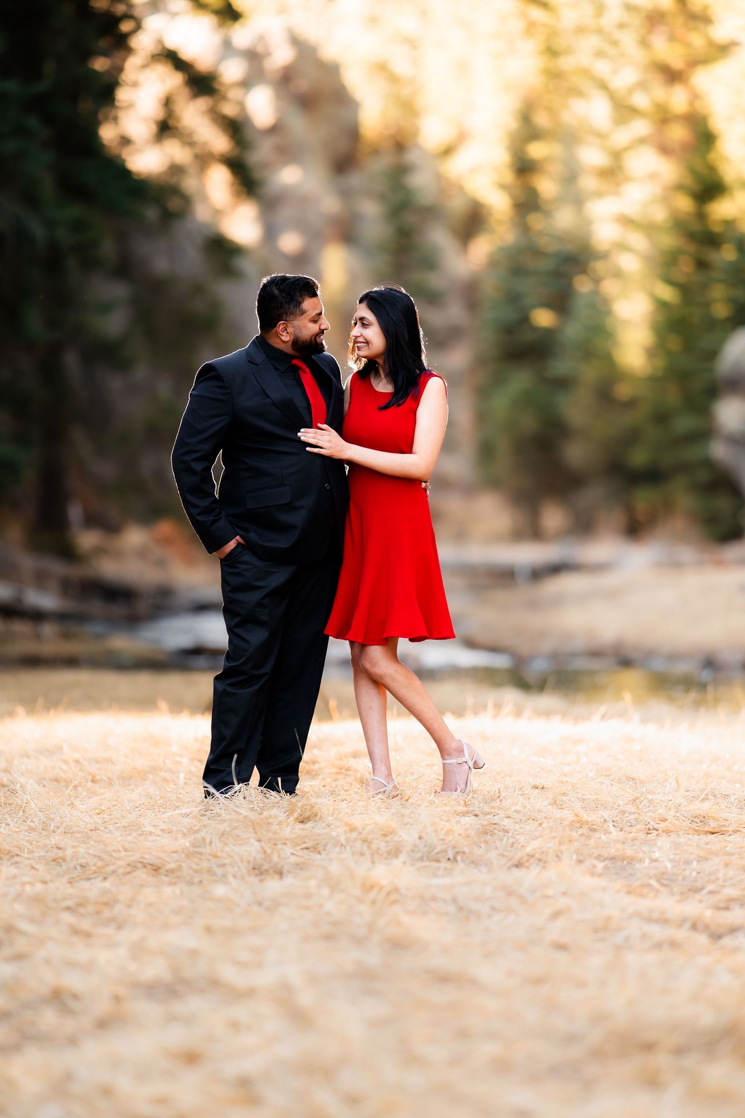 A couple stands together outdoors, with her wearing a red dress and him in black attire during golden hour.