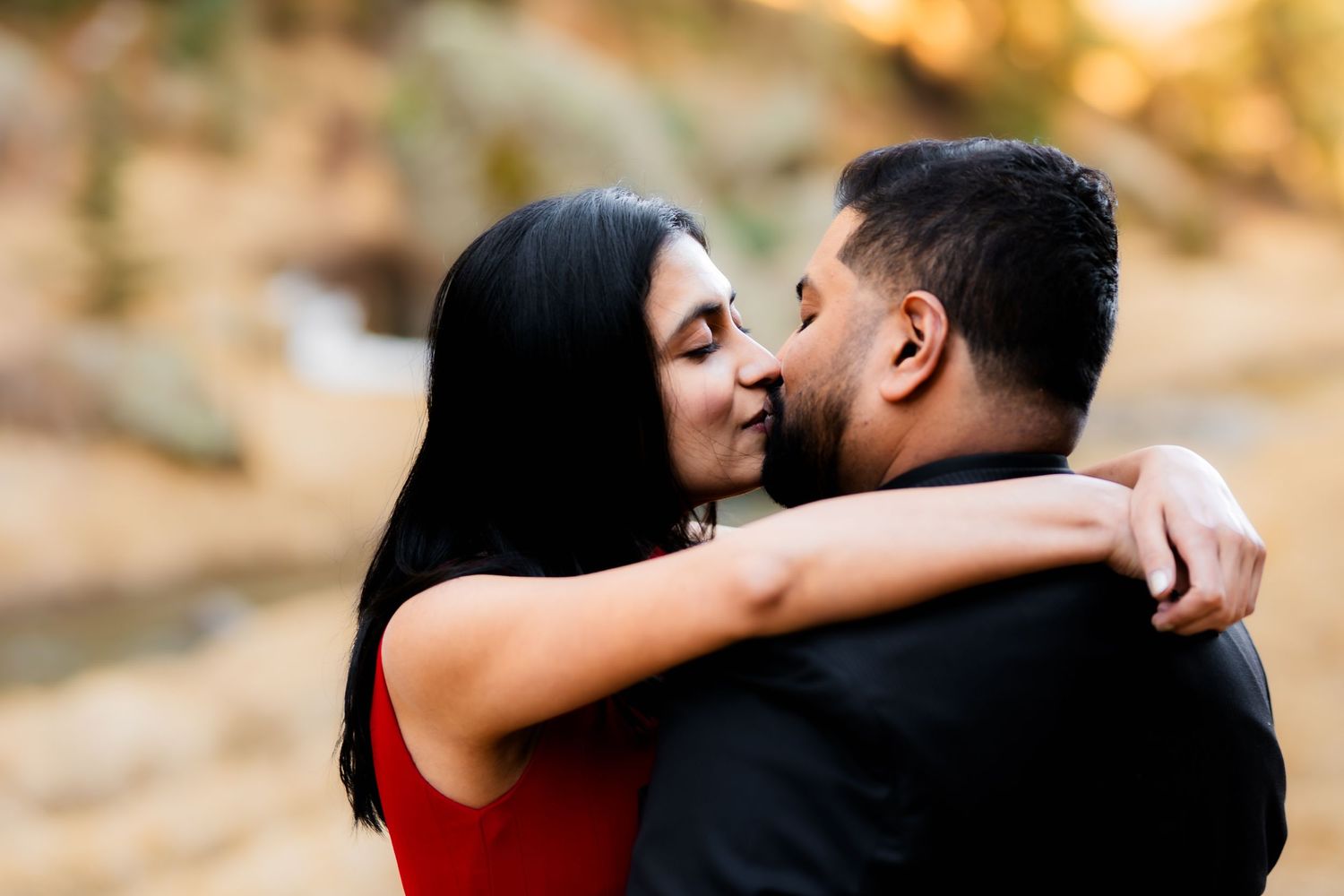 A couple sharing a romantic kiss outdoors during golden hour with sunlit trees in the background.