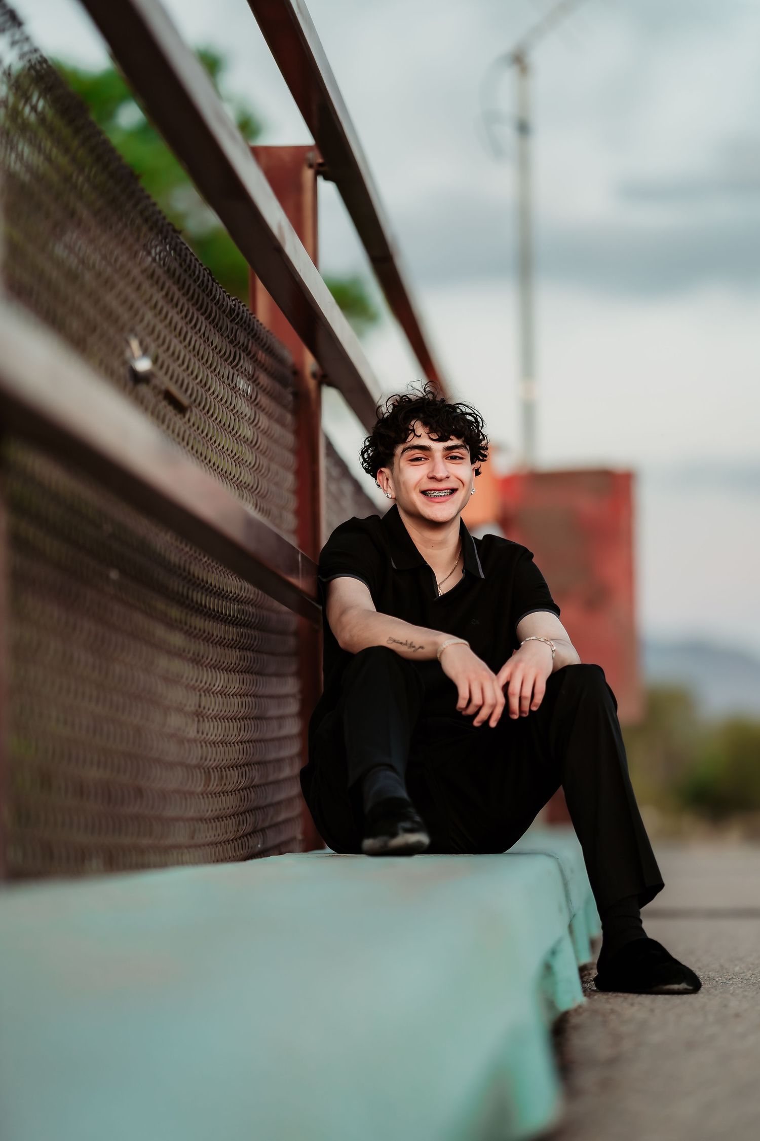 A person in all black casual wear poses while sitting on a concrete ledge against an urban backdrop on a cloudy day.