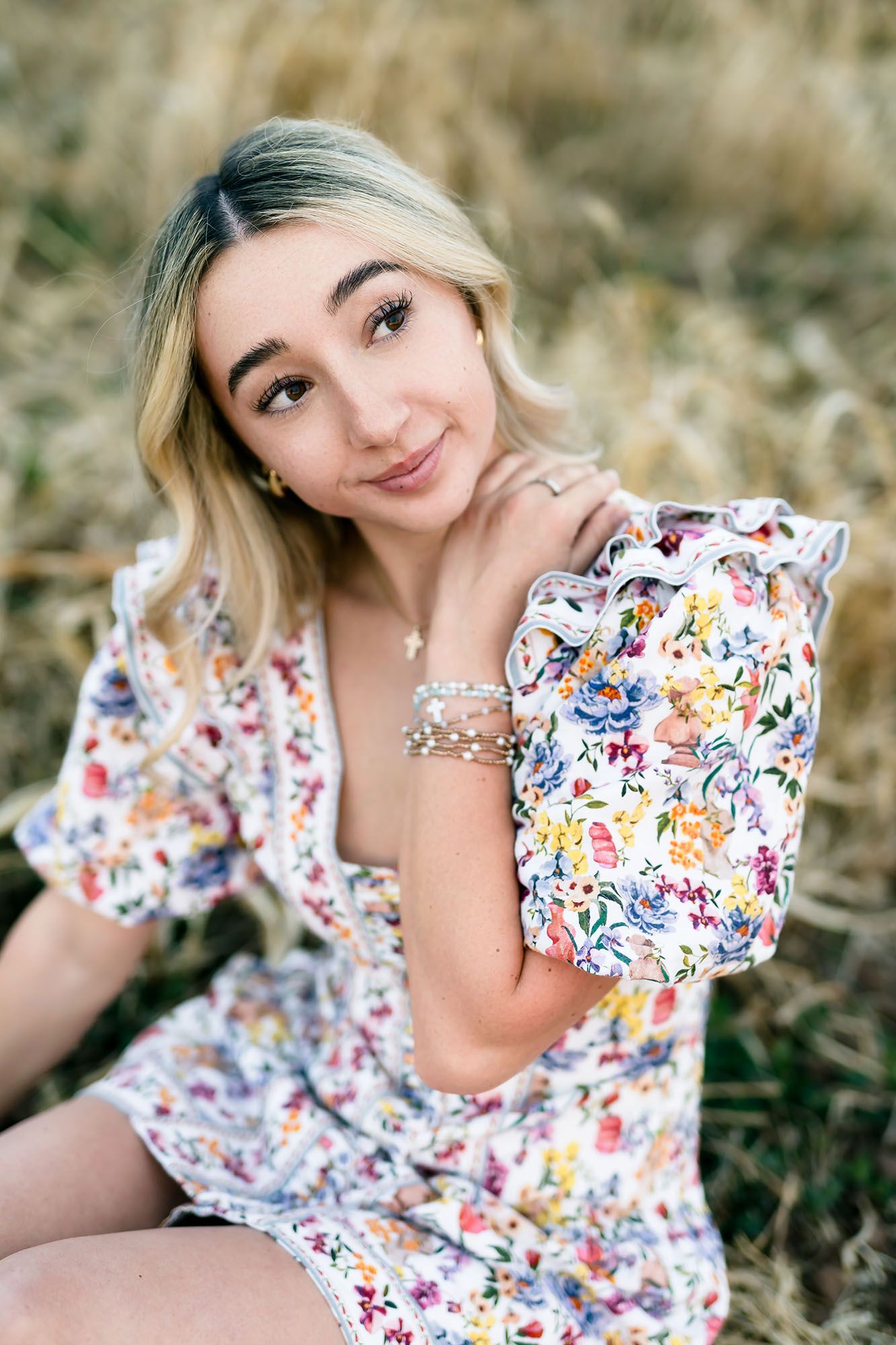 Young woman in floral dress sitting in a field, smiling softly while holding a ruffled floral bag.
