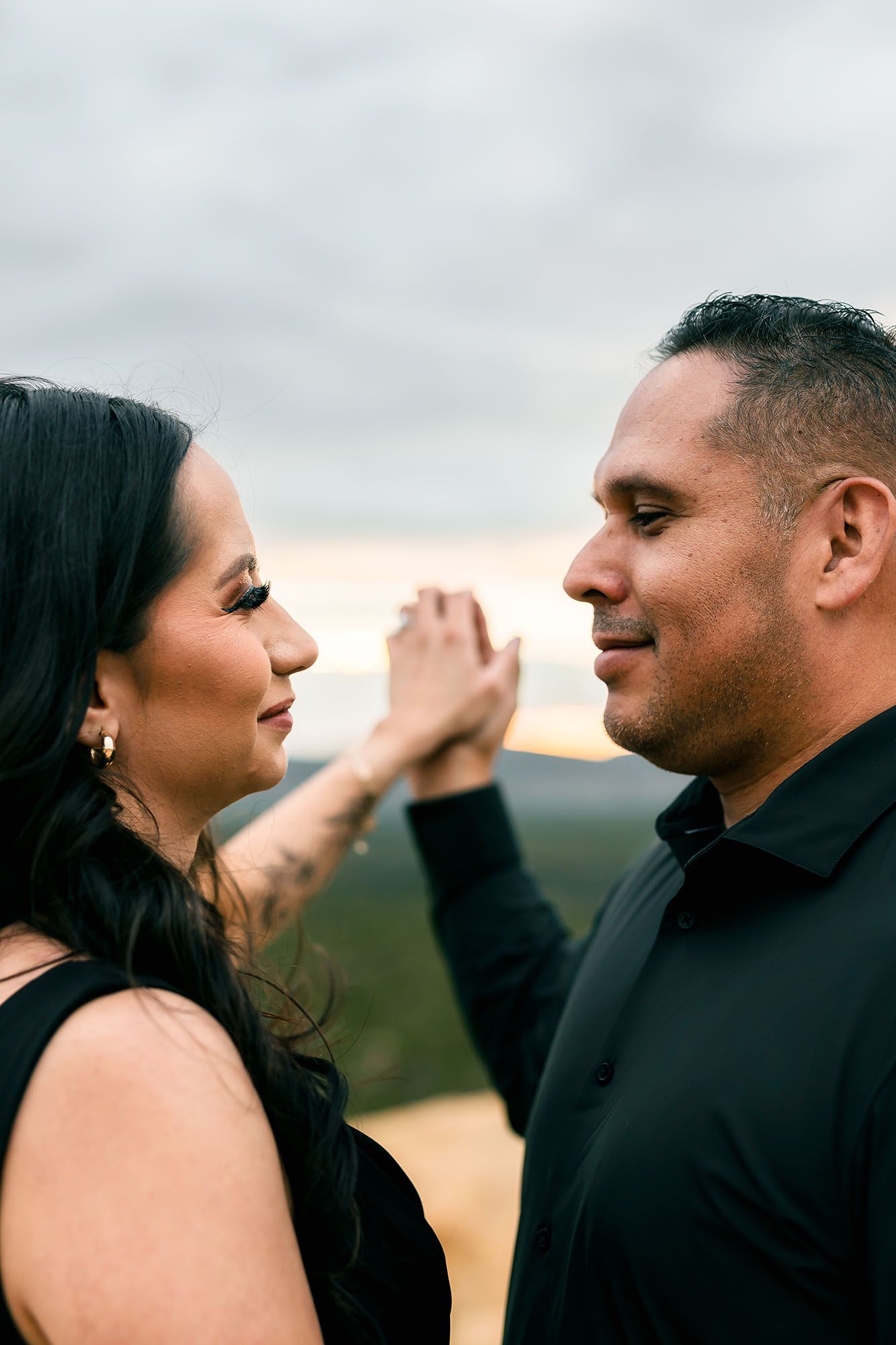 Couple in black outfits sharing a tender moment, woman gently touching man's face at sunset.