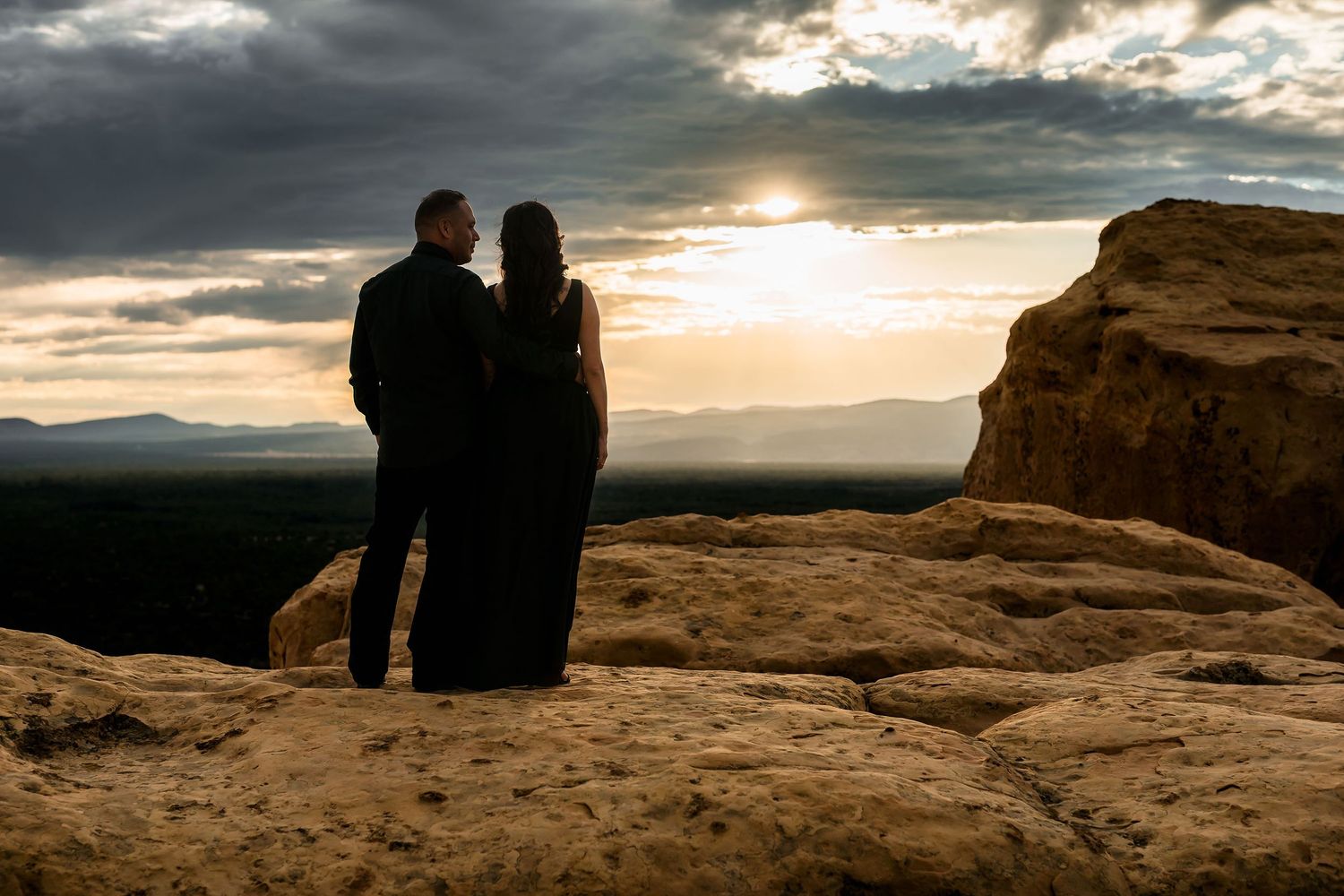 Couple in black attire sharing a romantic moment on desert rock at golden sunset.