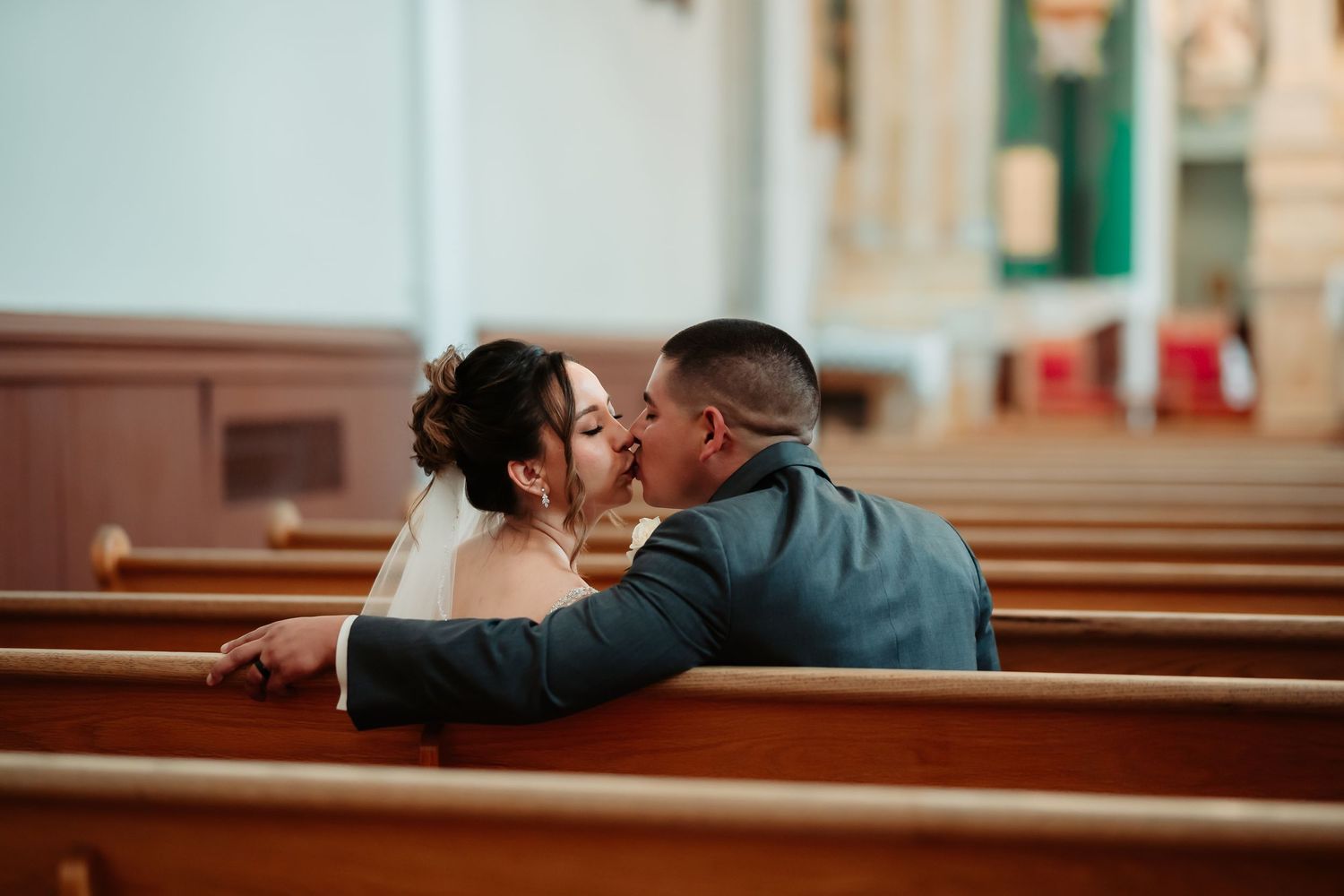 Couple kisses intimately sitting in wooden church pew with romantic lighting and warm tones.