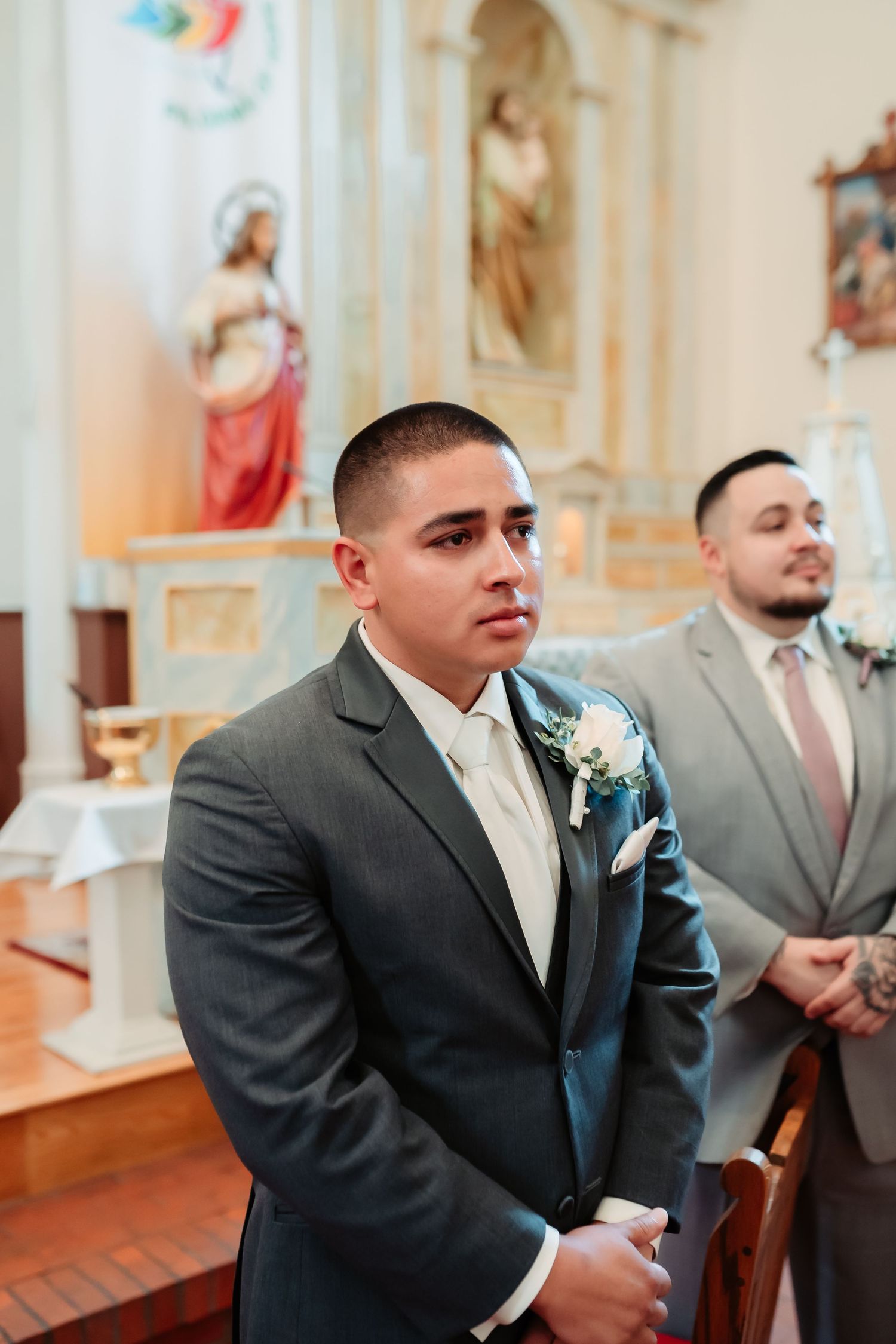 Groomsmen wearing gray suits and ivory ties seated in ornate church pews during wedding ceremony.