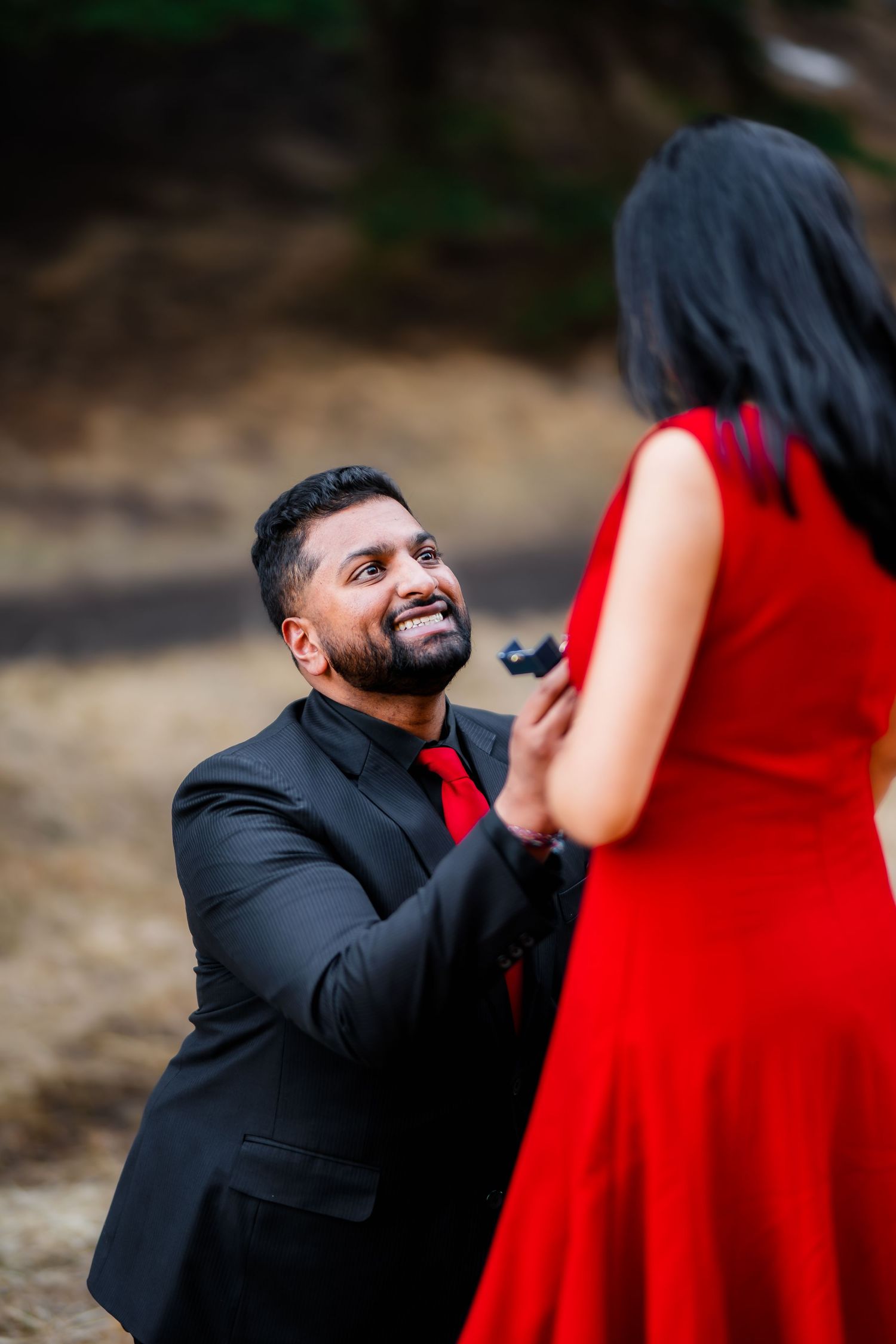 A proposal moment captured outdoors with a couple dressed in formal red and black attire.