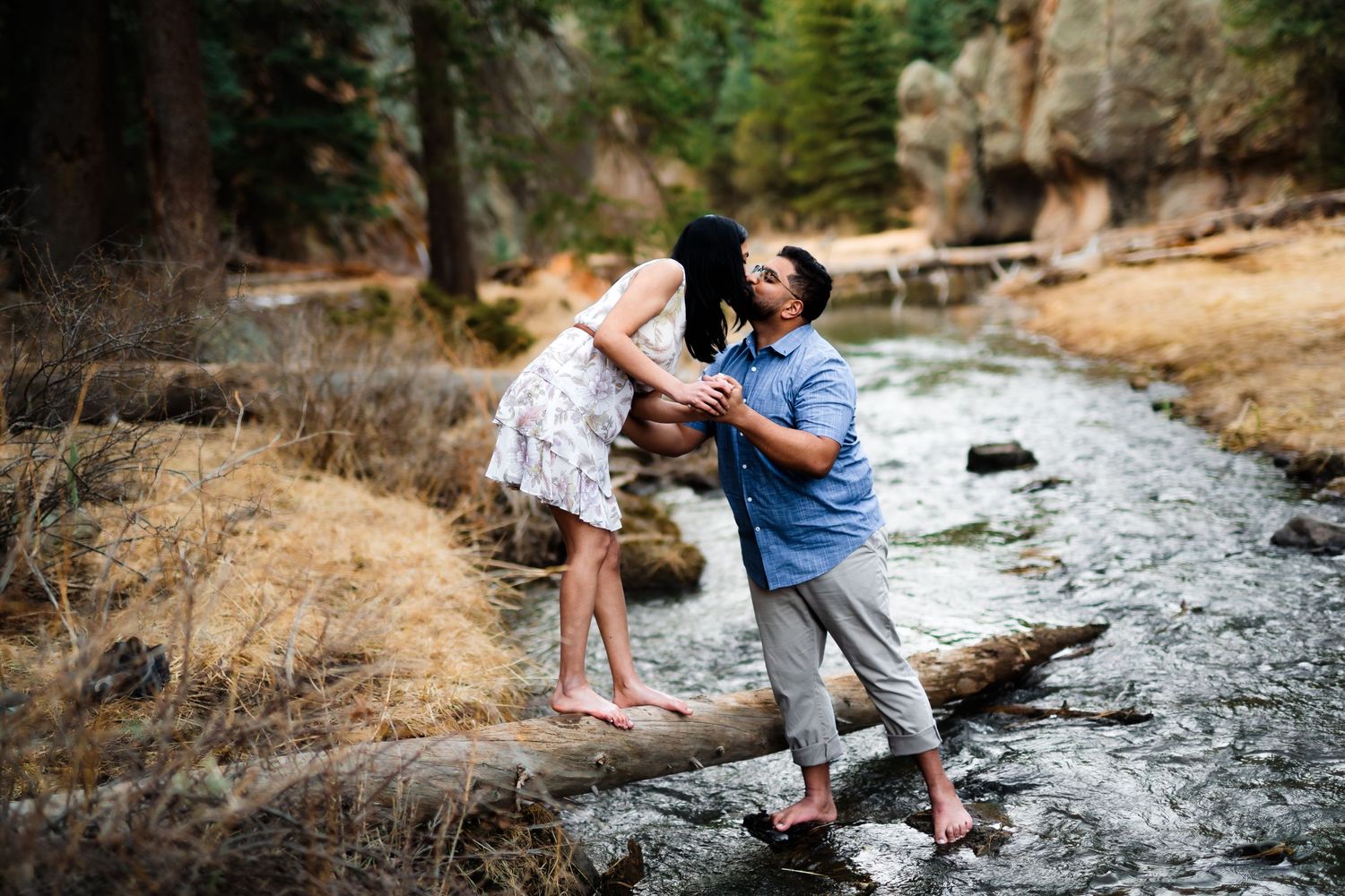 A playful couple balances on rocks while crossing a stream in a forest setting.