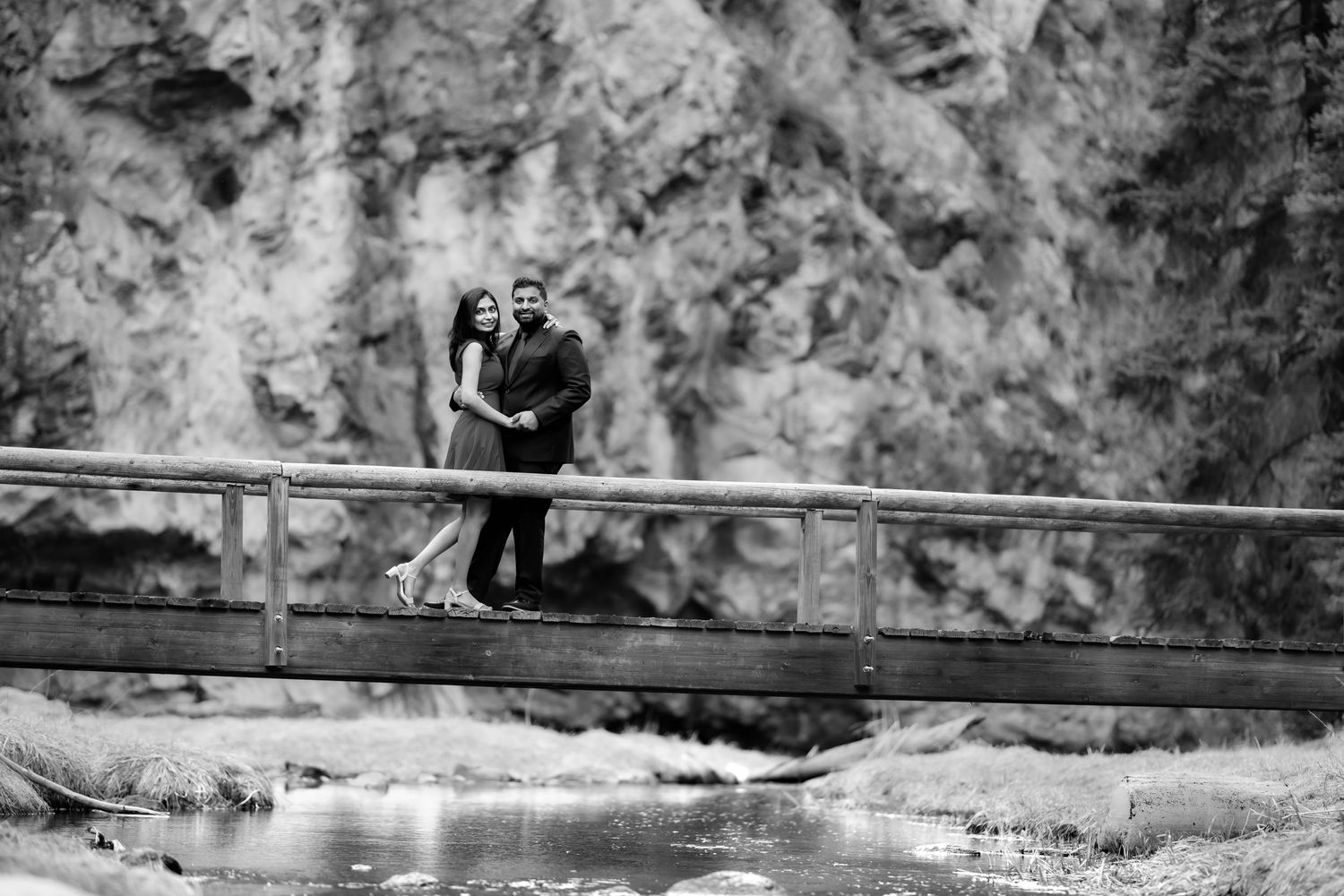 A couple stands together on a wooden bridge over water with rocky cliffs in the background.