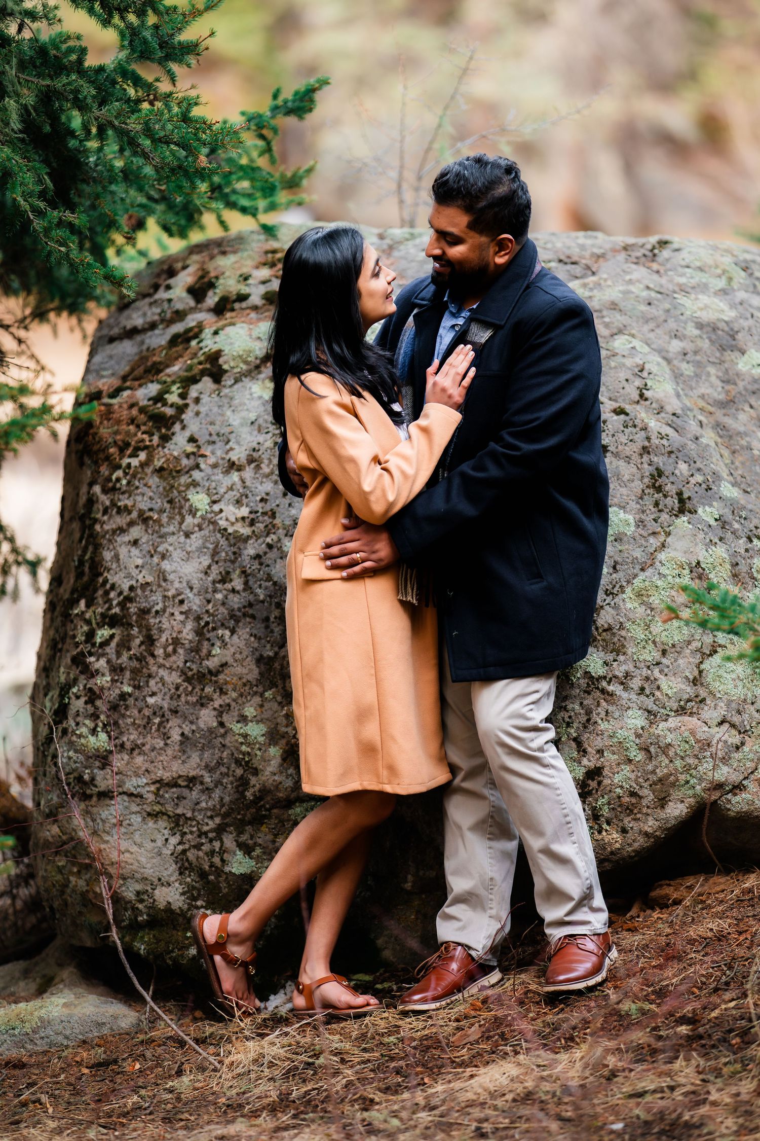 Romantic couple embraces against large boulder in outdoor setting with woman in tan coat and man in black jacket.