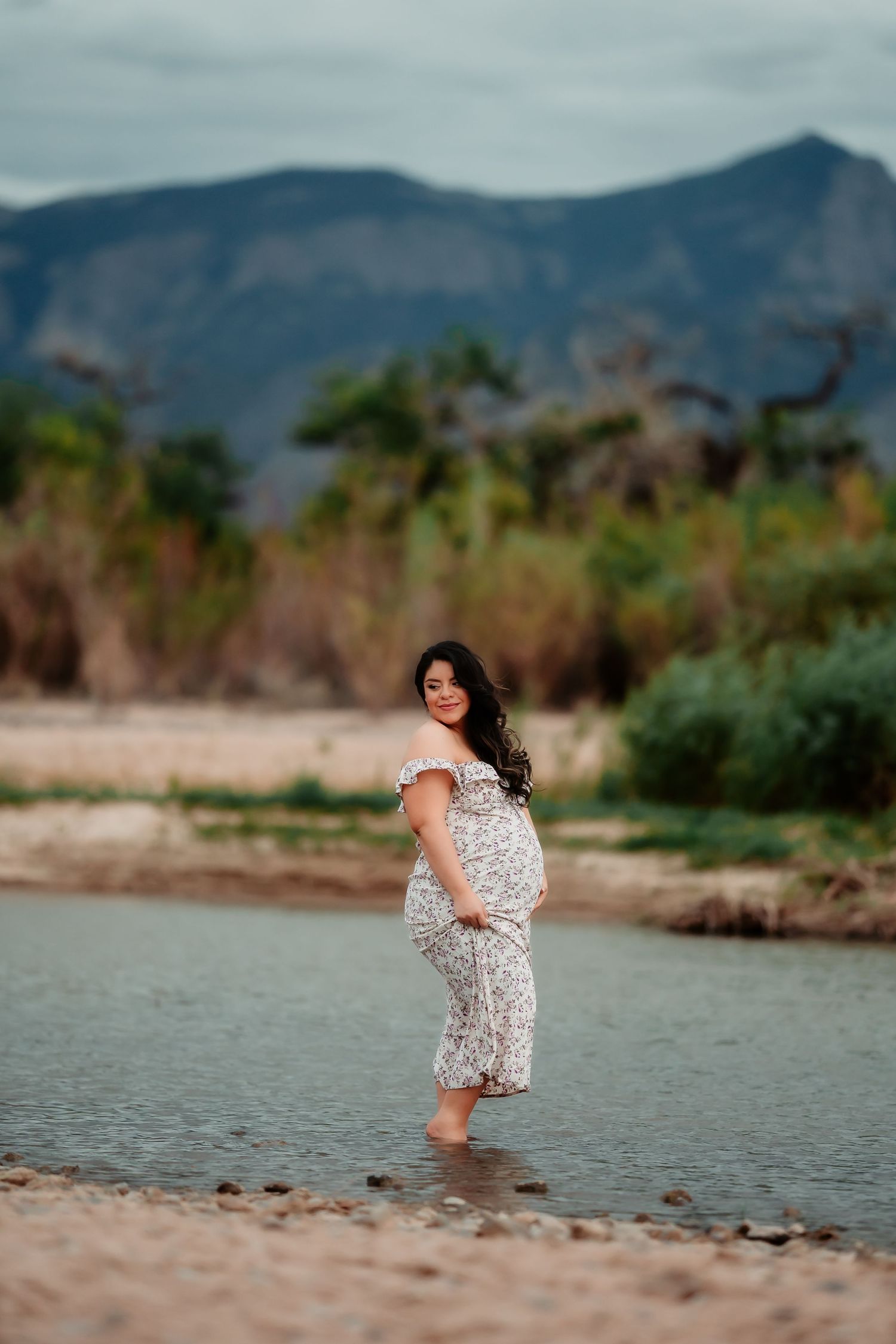 A maternity photoshoot by a lake with mountains in the background showing playful poses.