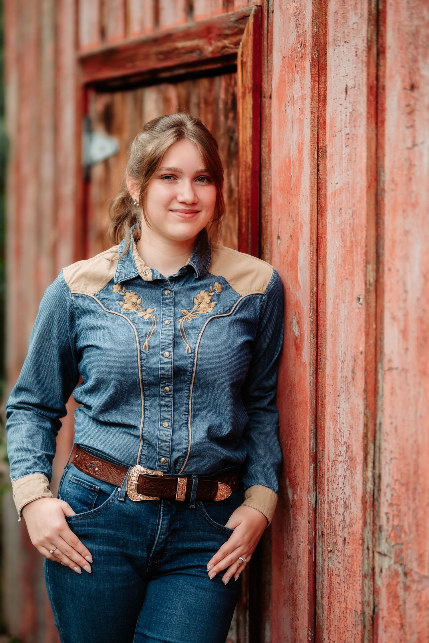 Person wearing denim jacket and jeans poses against rustic red barn wall in country fashion photoshoot.