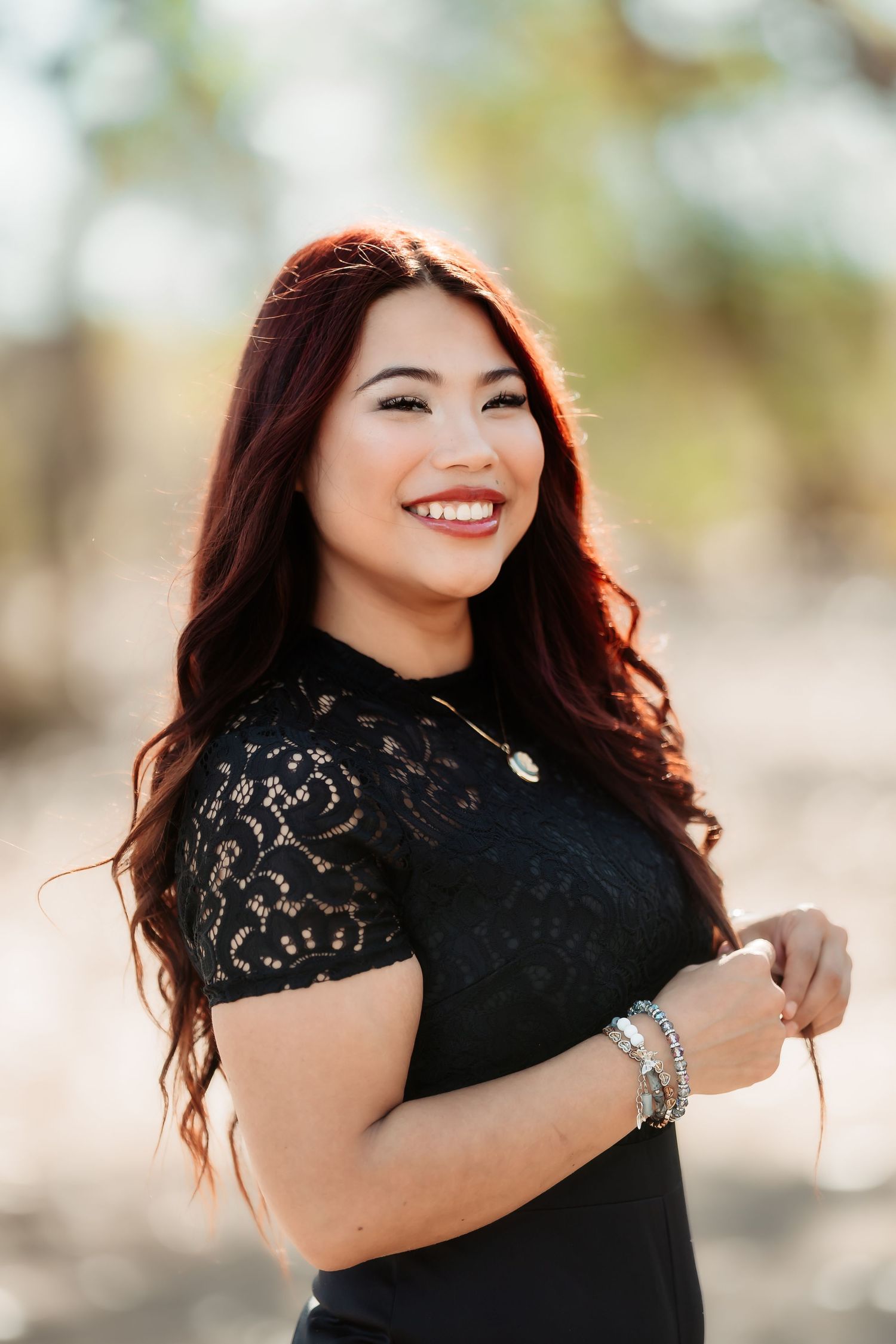 Woman in black lace top smiles radiantly while posing outdoors in natural lighting.