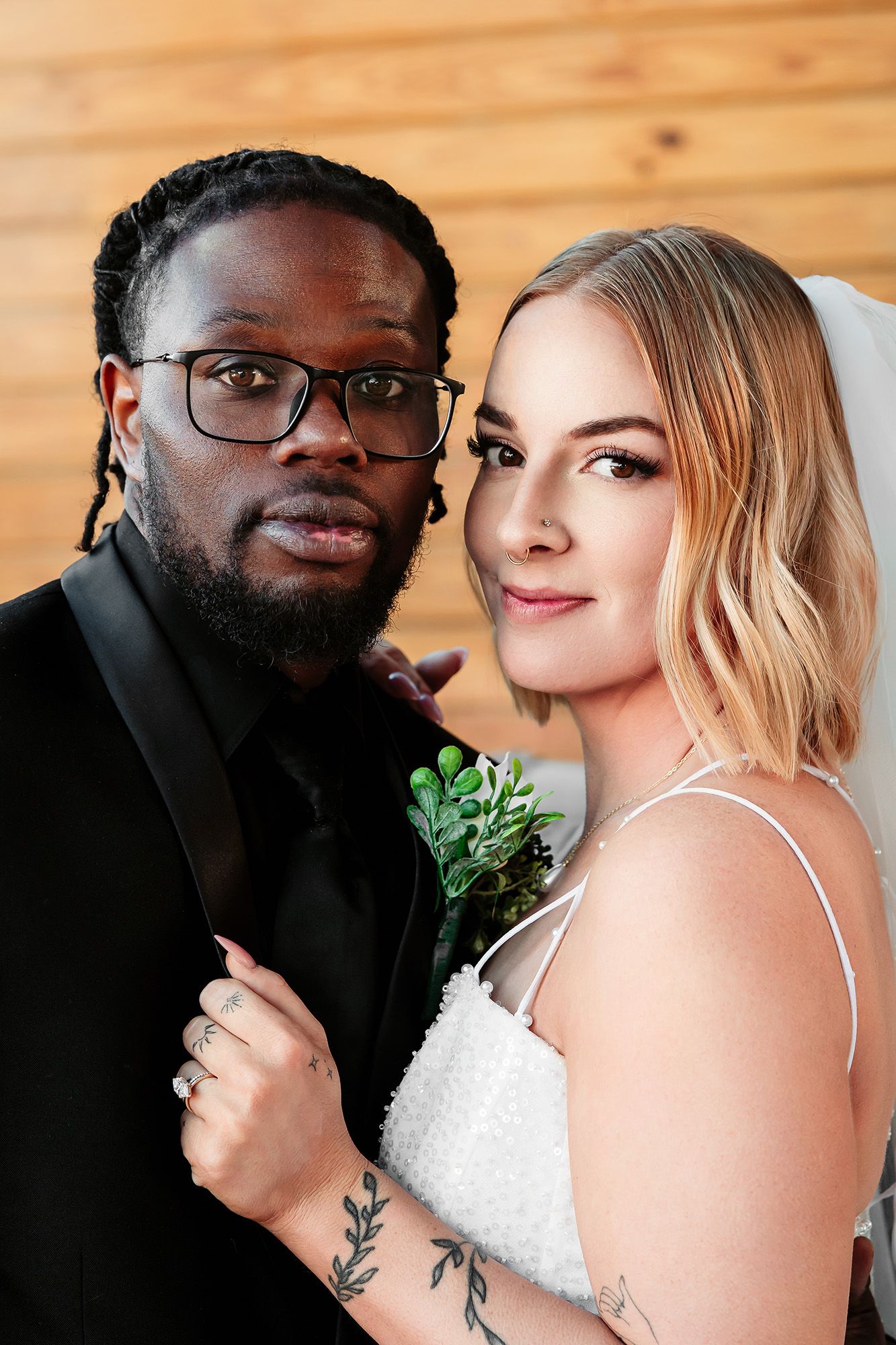 Interracial couple in wedding attire pose closely, groom in black suit with boutonniere, bride in white dress with veil.