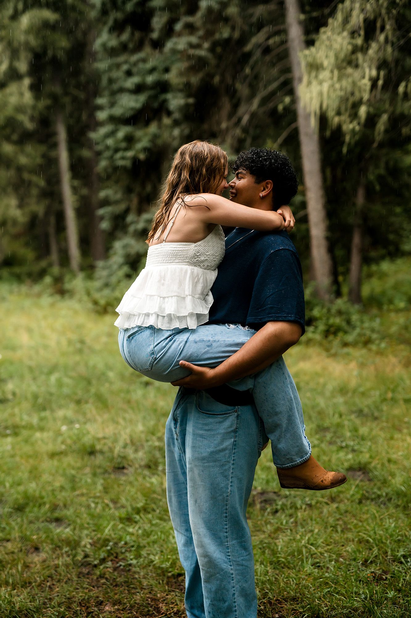 A man lifts a woman in his arms outdoors, sharing a romantic moment in a lush green forest setting.