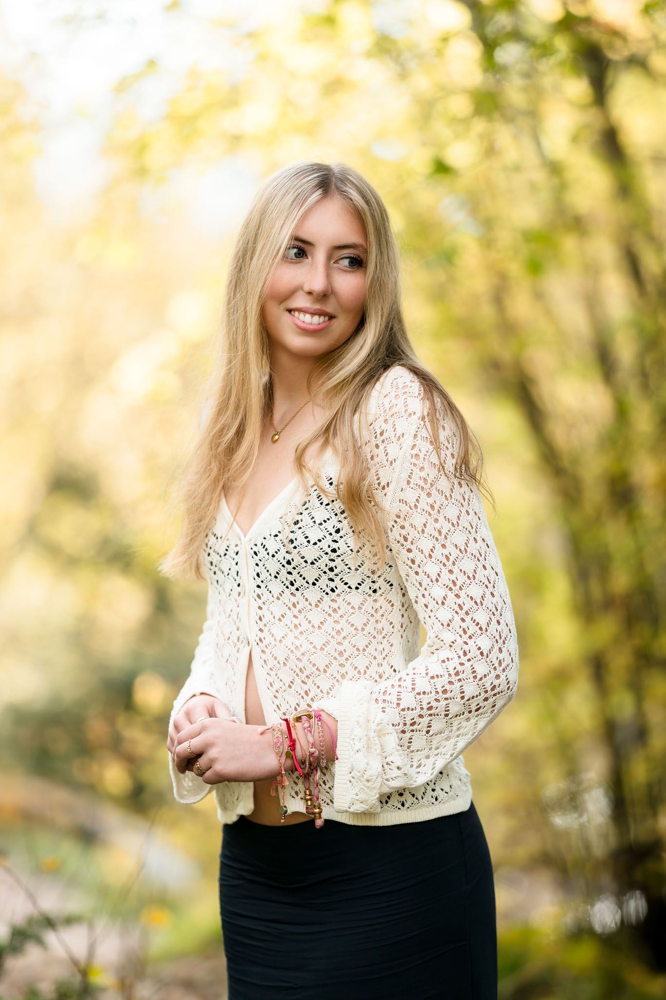 Smiling blonde woman in white crochet cardigan and black skirt posing outdoors among autumn foliage.