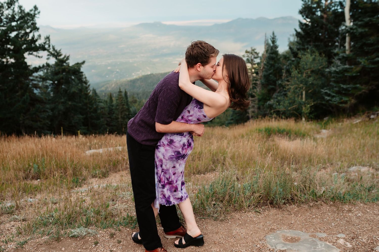 A couple embraces on a mountainside with distant hills in view.