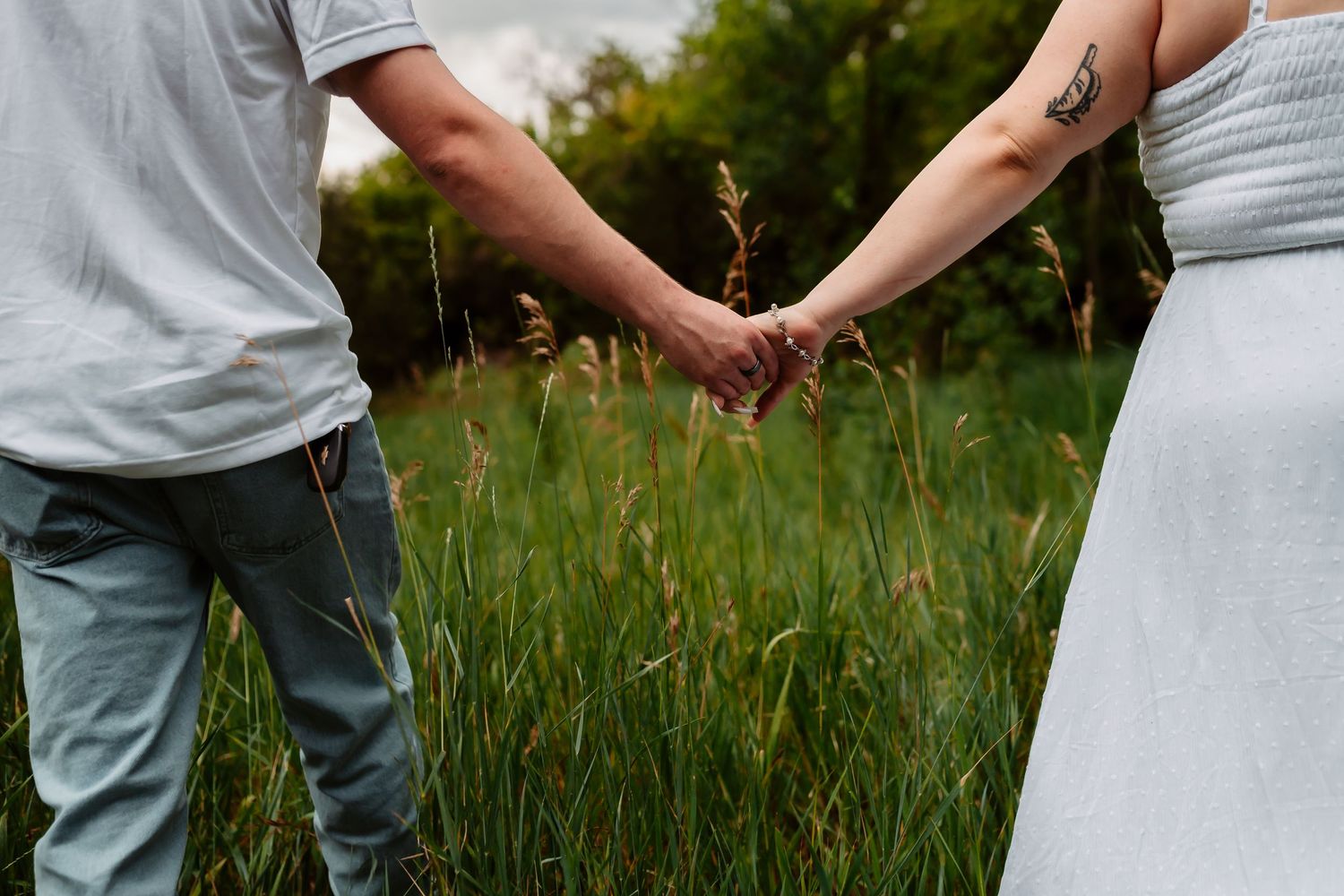 A couple in grey and white outfits holding hands in a field of grass during sunset.