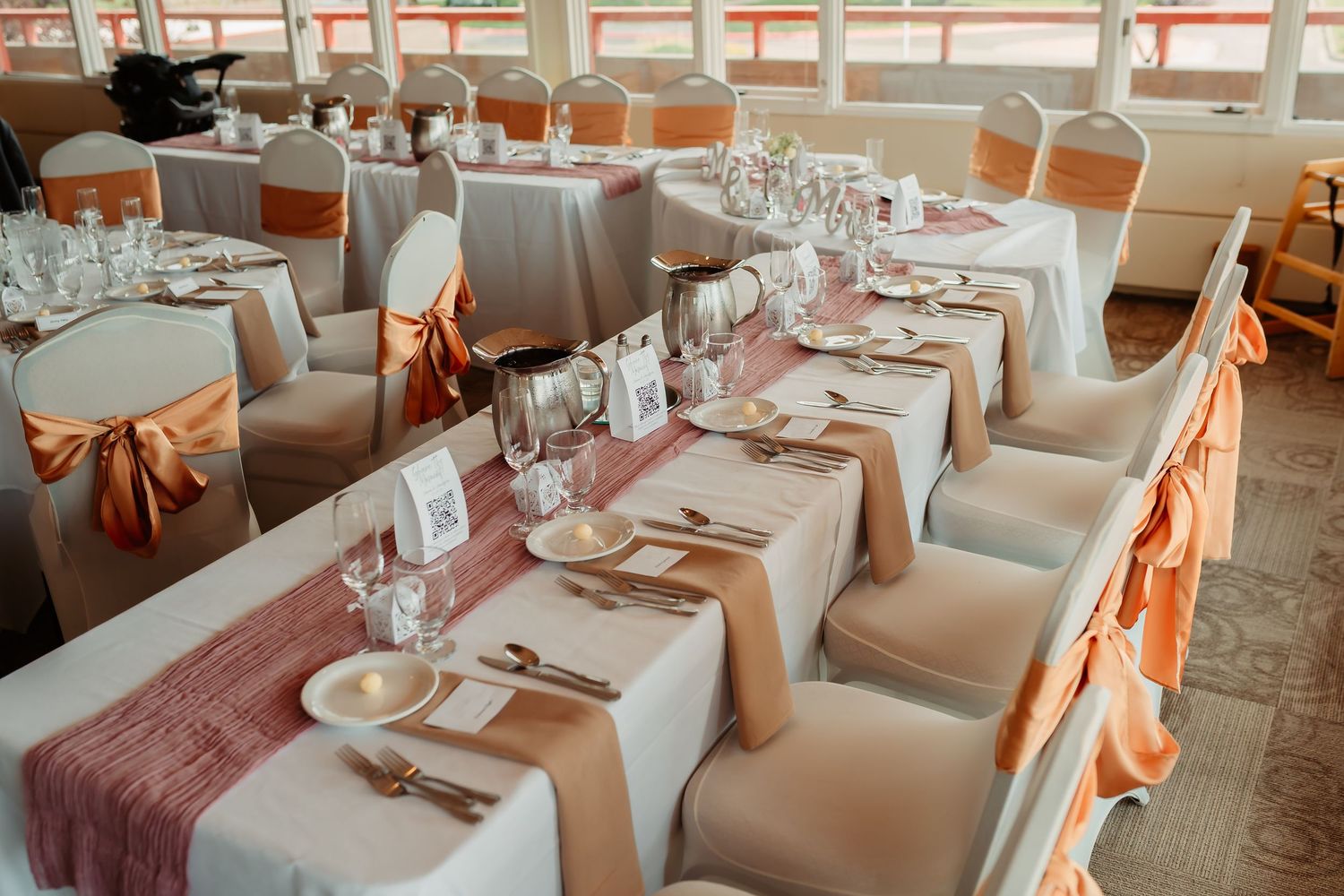 An elegant wedding reception venue with round tables, orange satin chair sashes, and romantic lighting creates a warm atmosphere.