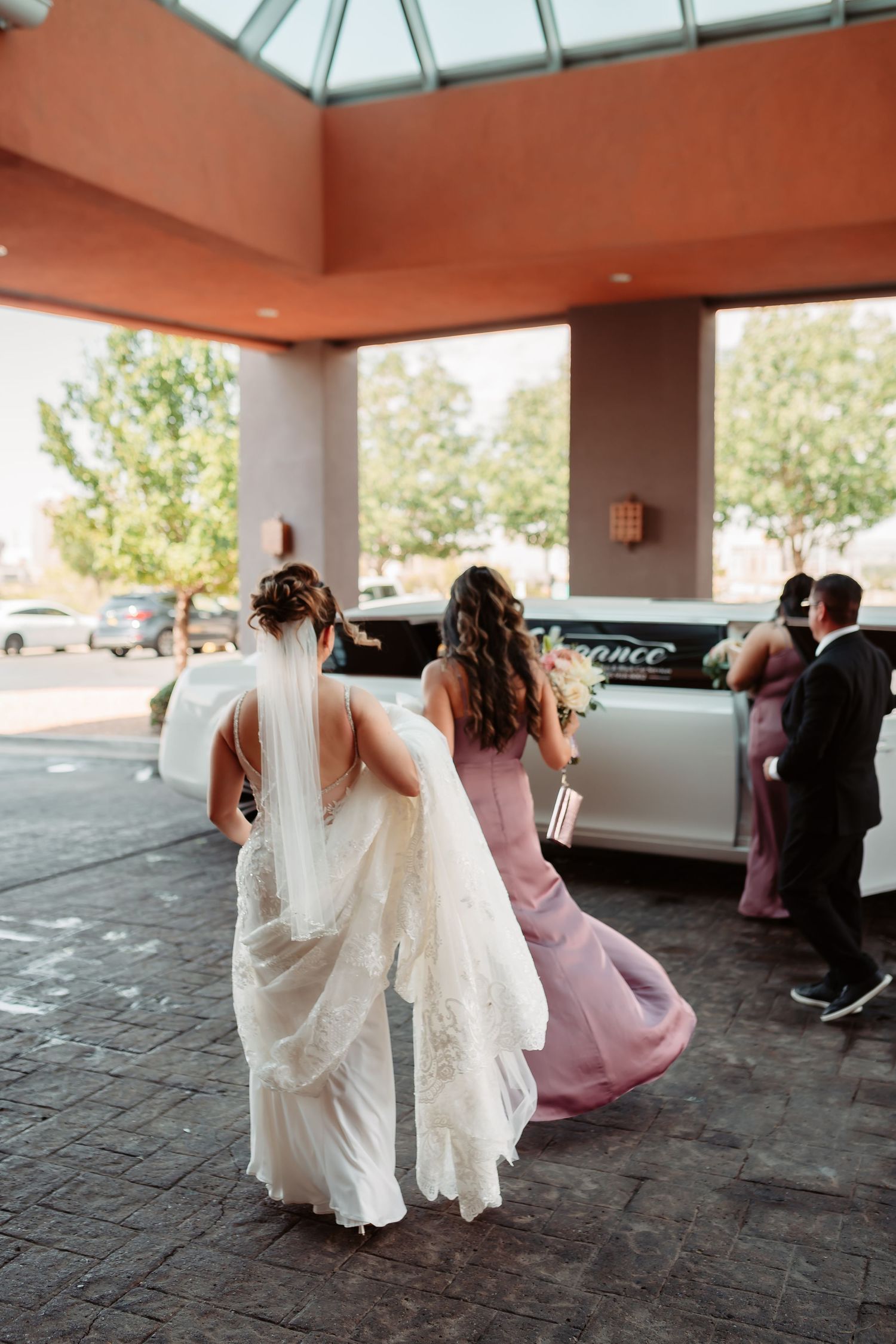 A bridal party runs playfully through a covered parking area, with the bride's white dress and bridesmaids' pink gowns flowing.