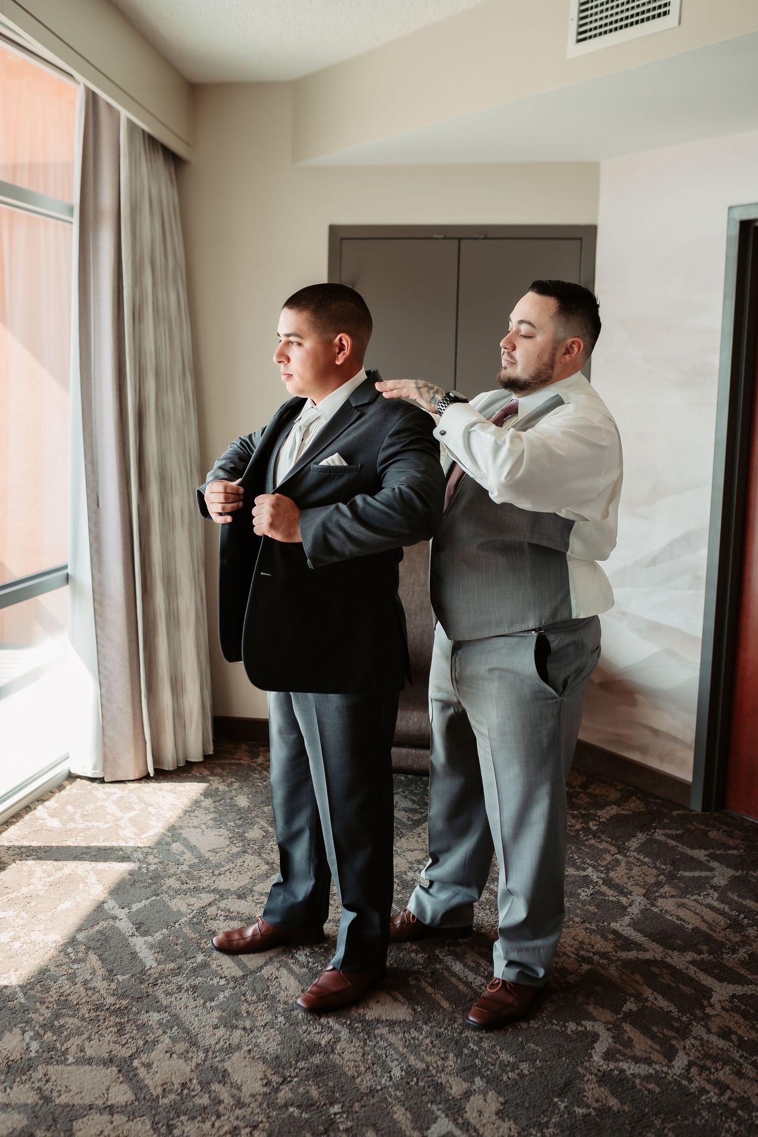 Men in formal suits helping each other get dressed in a hotel room with patterned carpet.