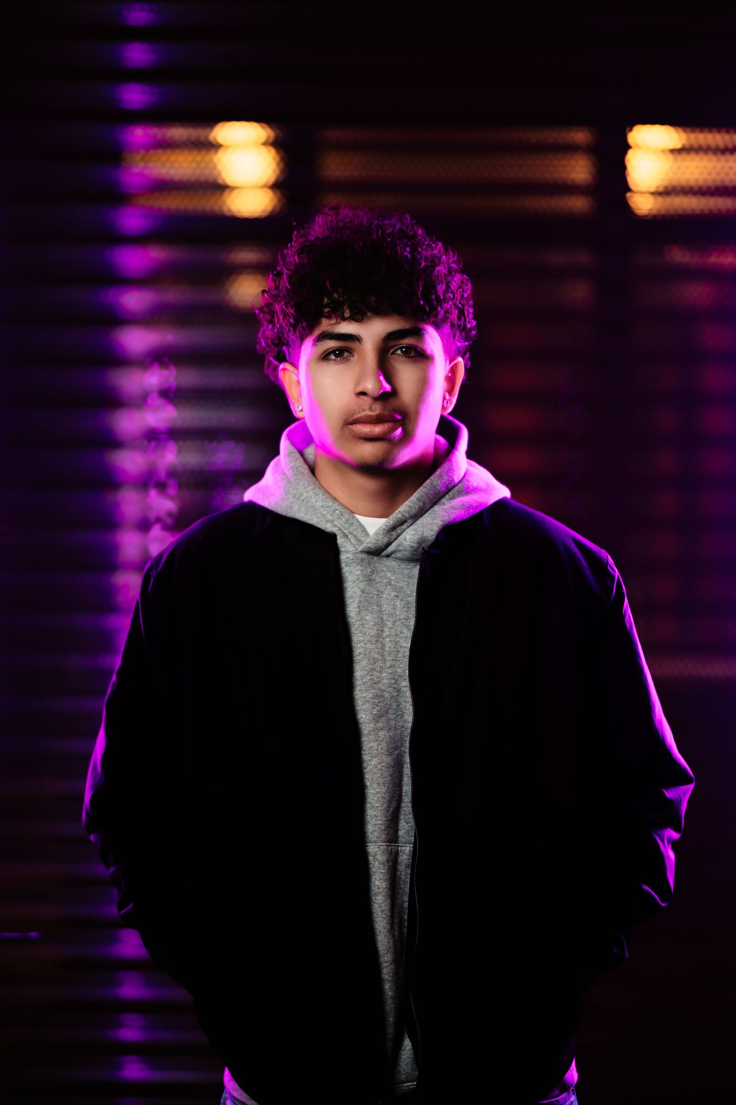 A person in a black jacket and gray hoodie poses against a dark background with purple lighting effects.