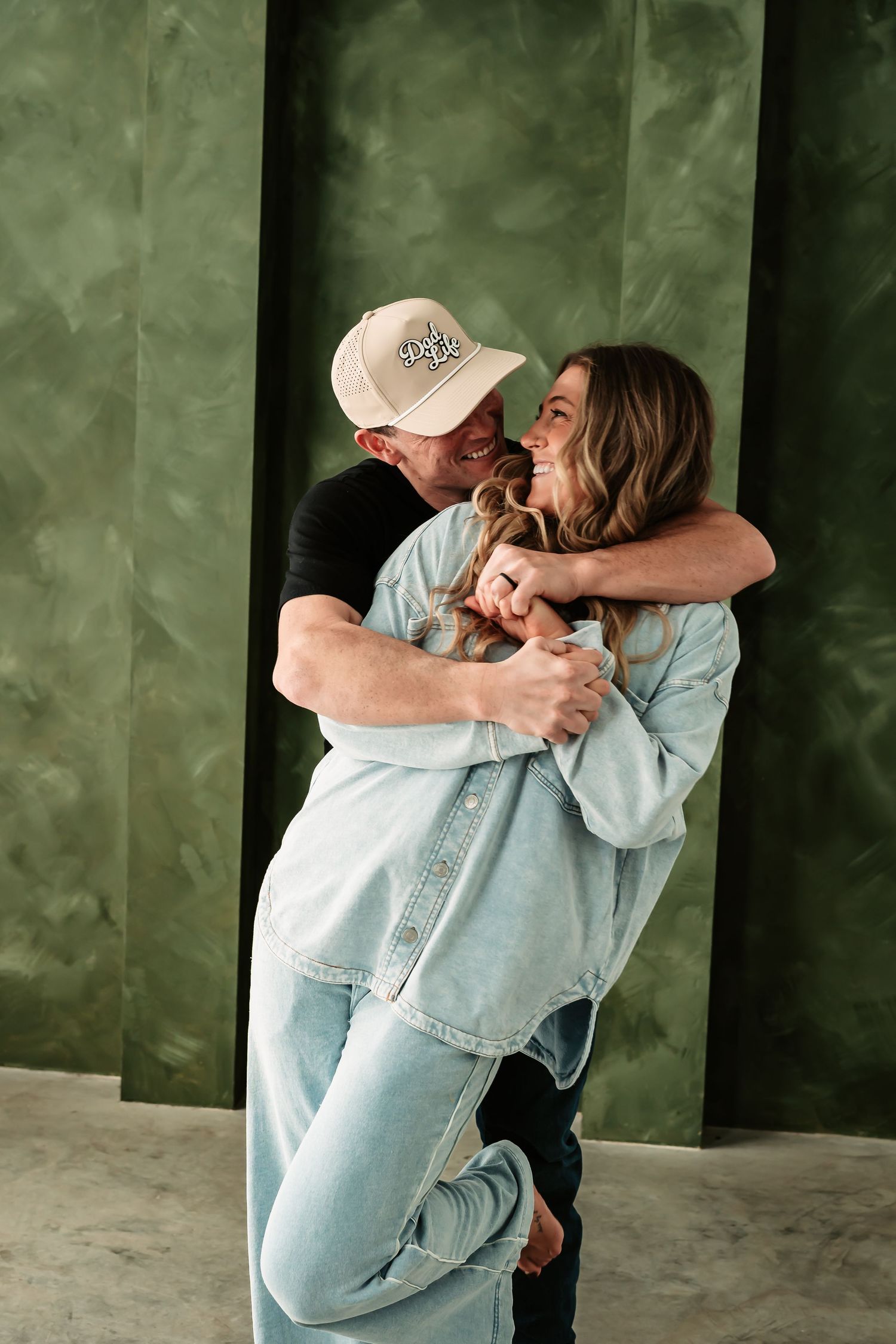 A couple embraces and laughs together against a green textured wall, wearing casual denim and a baseball cap.