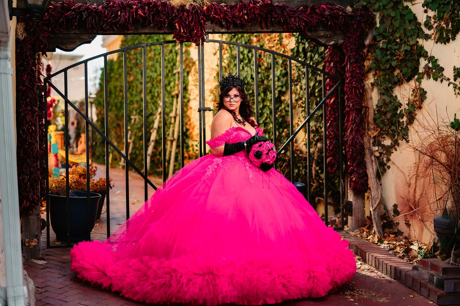 A person wearing a vibrant hot pink tulle ballgown poses in a garden setting with wrought iron gates.