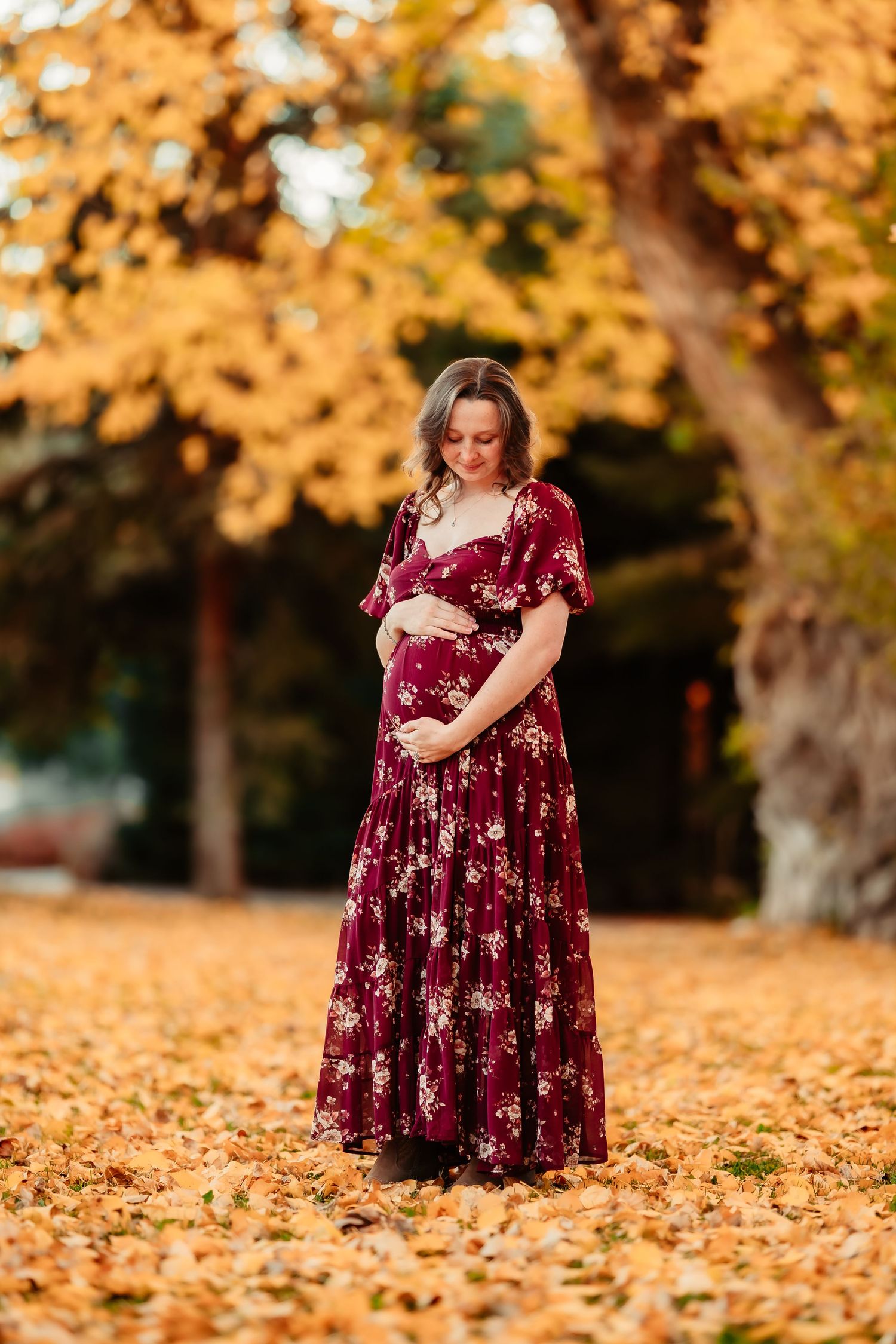 A woman in a burgundy floral dress stands among golden autumn leaves in a park setting.