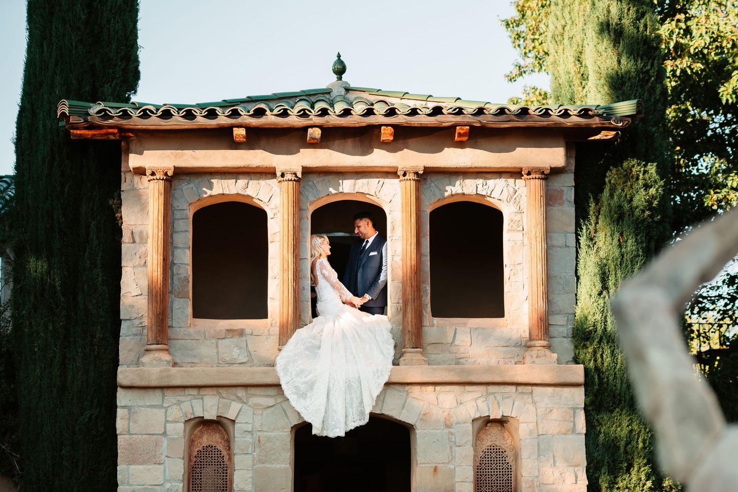 A couple poses in front of a stone building with terracotta roof tiles during a sunny outdoor wedding photoshoot.