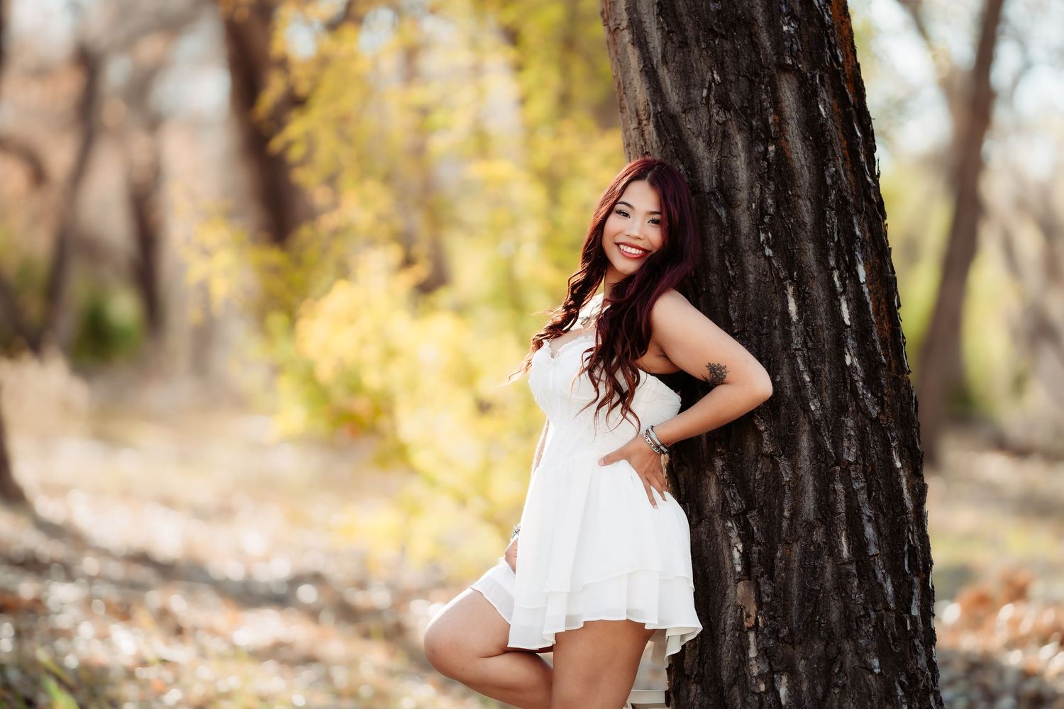 A person in a white dress poses gracefully against a tree trunk in an autumn forest setting.