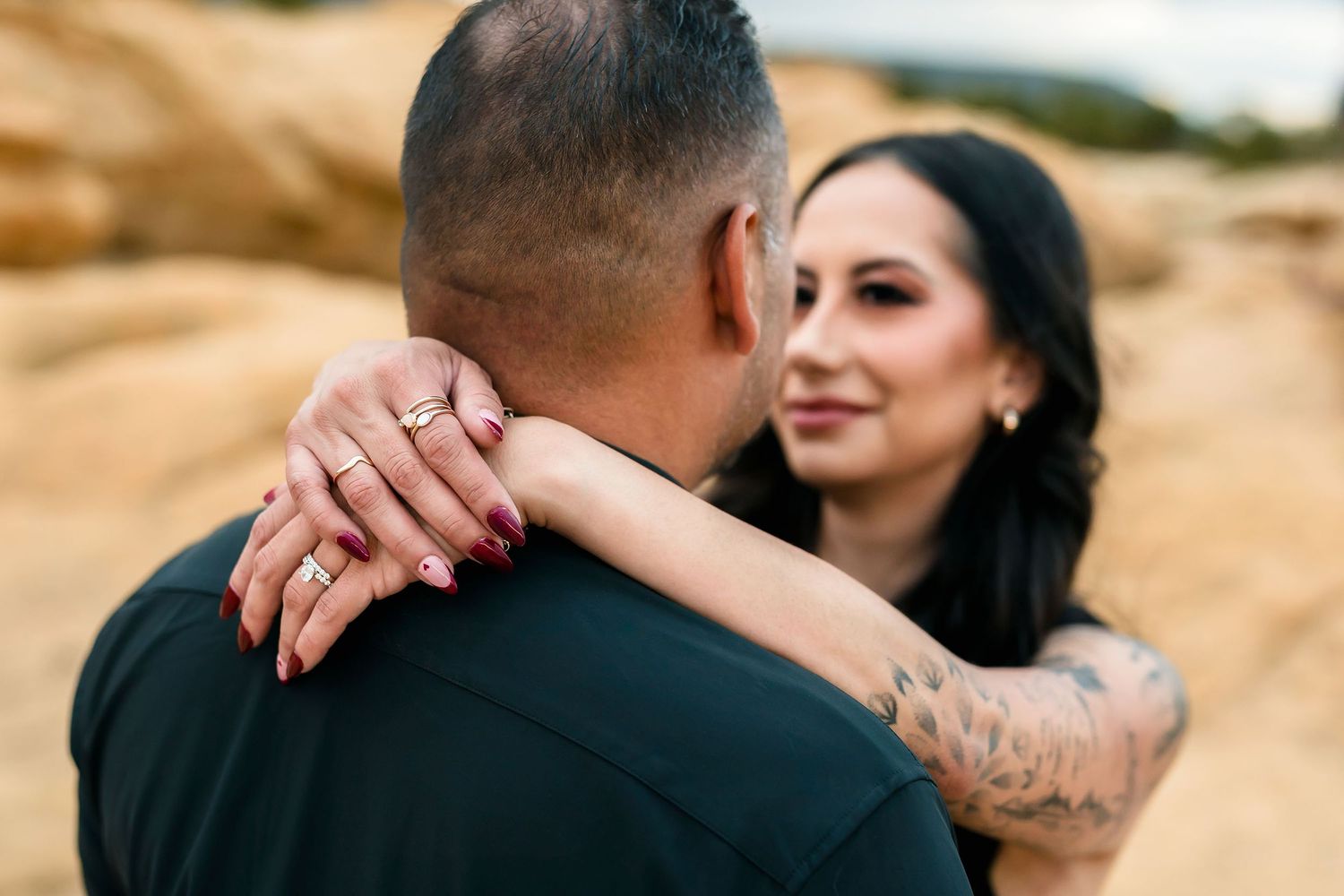 Close-up of couple embracing on rocky terrain, woman with tattoos and gold rings resting hand on mans shoulder.